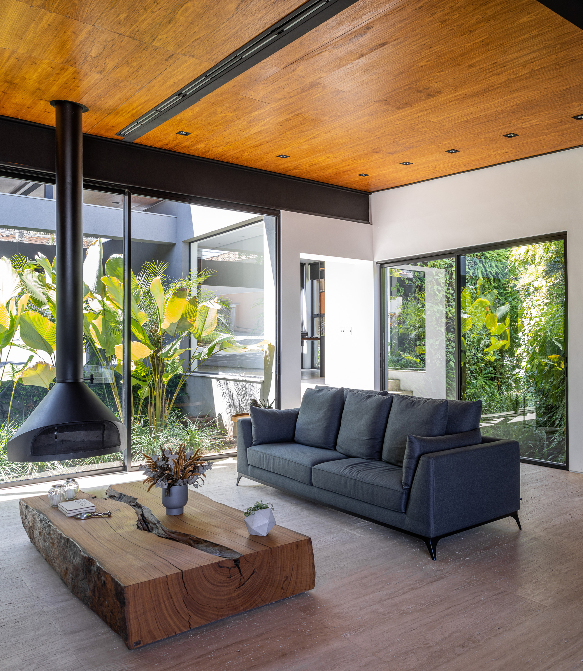 Living room with wood ceiling, suspended fireplace and glazed walls opening to planted courtyards