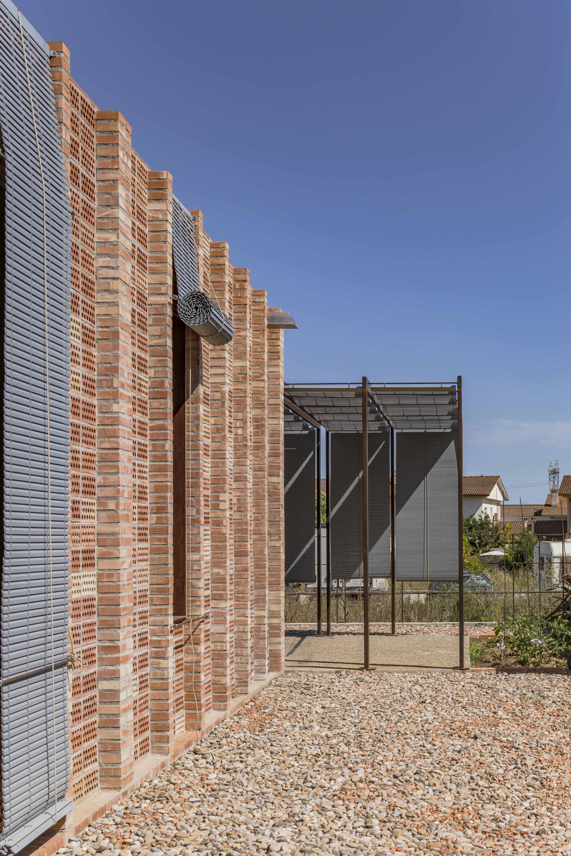 Perforated brick wall with horizontal louvers beside a metal-framed pergola on gravel ground