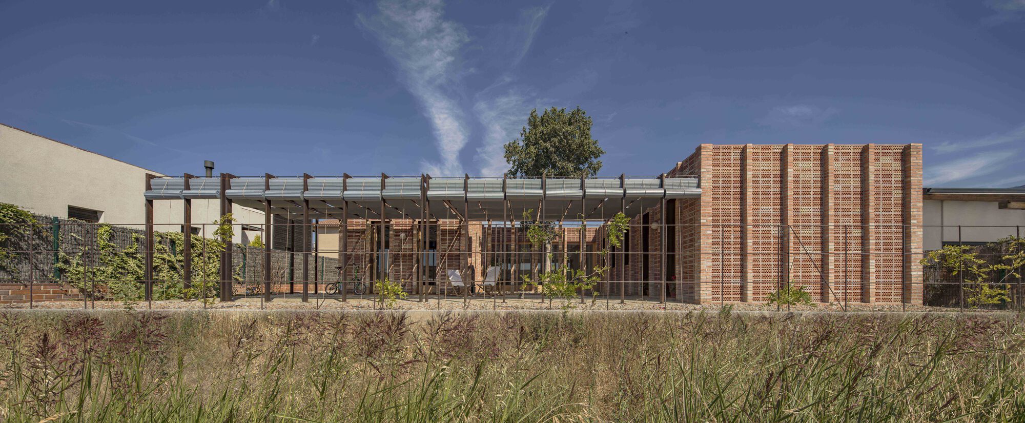 Street elevation showing perforated brick facade with timber pergola and native grasses in foreground under clear sky