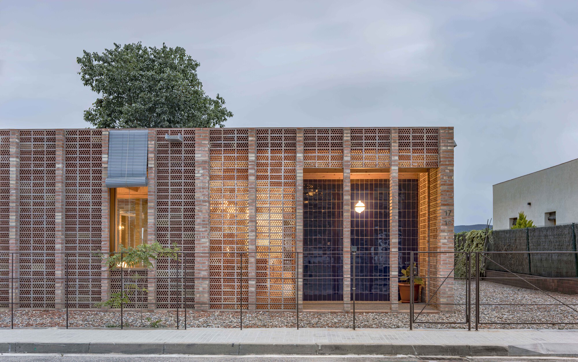 Street facade at dusk showing illuminated interior through perforated brick and steel mesh screens with tree above