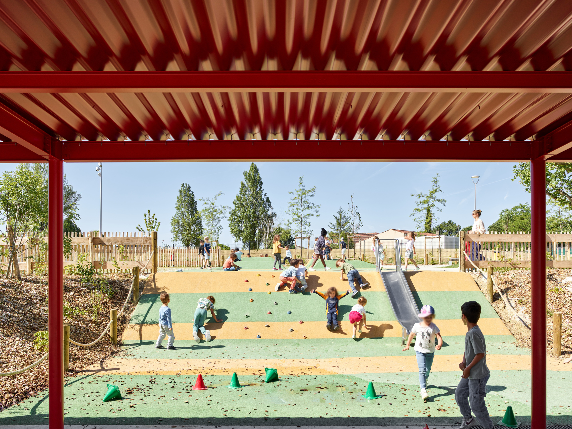 Red steel canopy framing a playground with children on a climbing hill and scattered play equipment