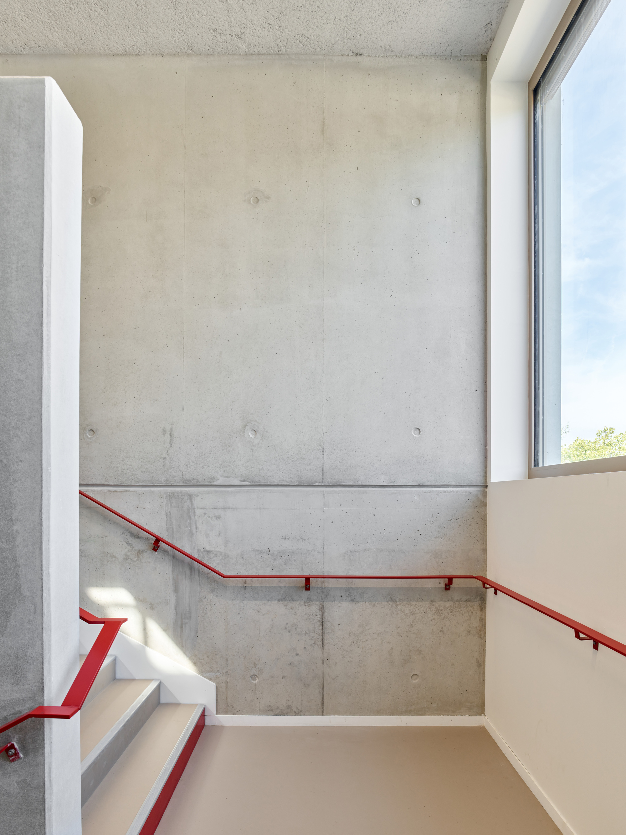 Stairwell with cast concrete walls, red steel handrail and natural light from a corner window
