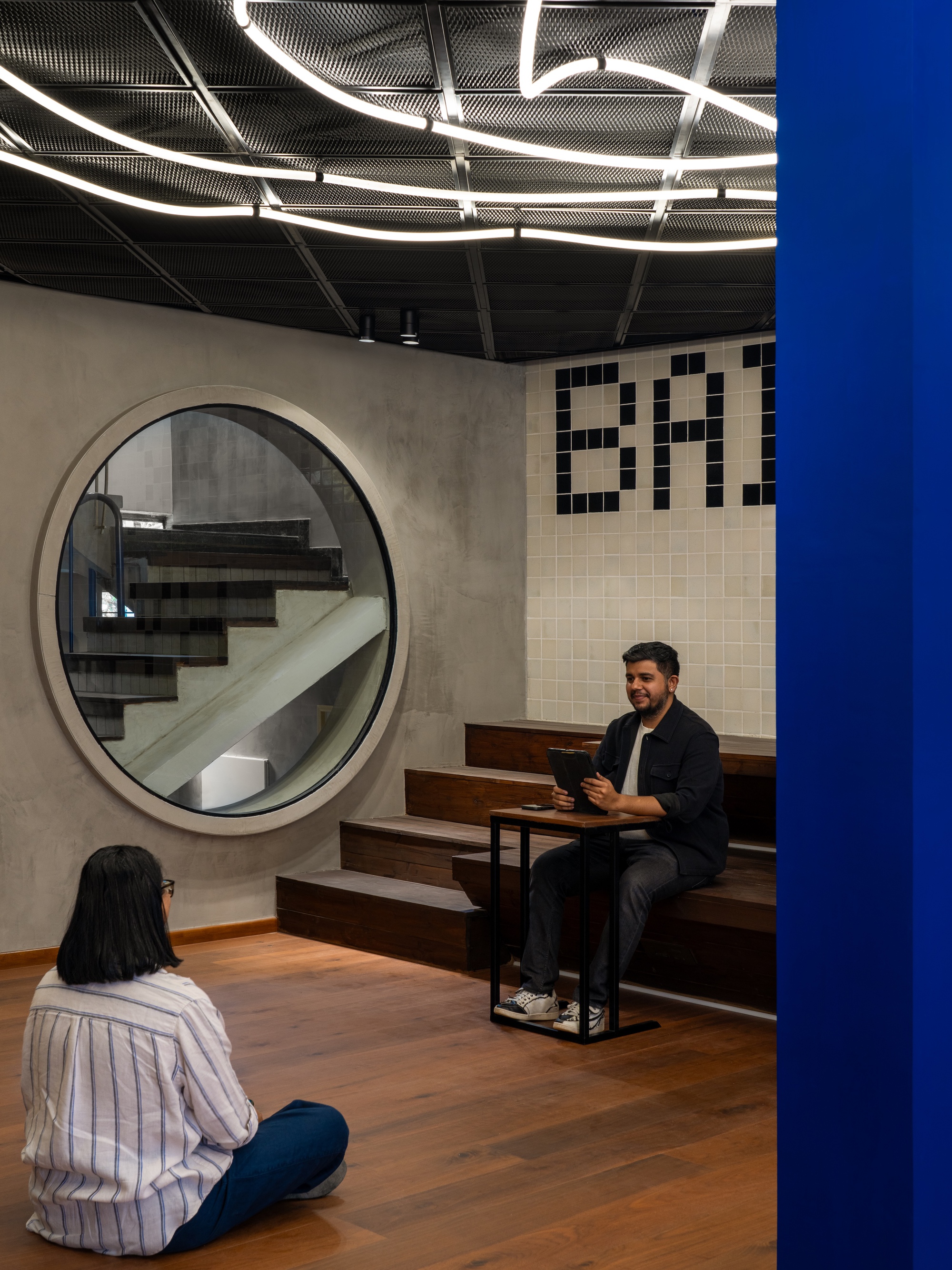 Seating area with circular concrete staircase portal, tiled wall graphics, and two people beneath curved linear lighting