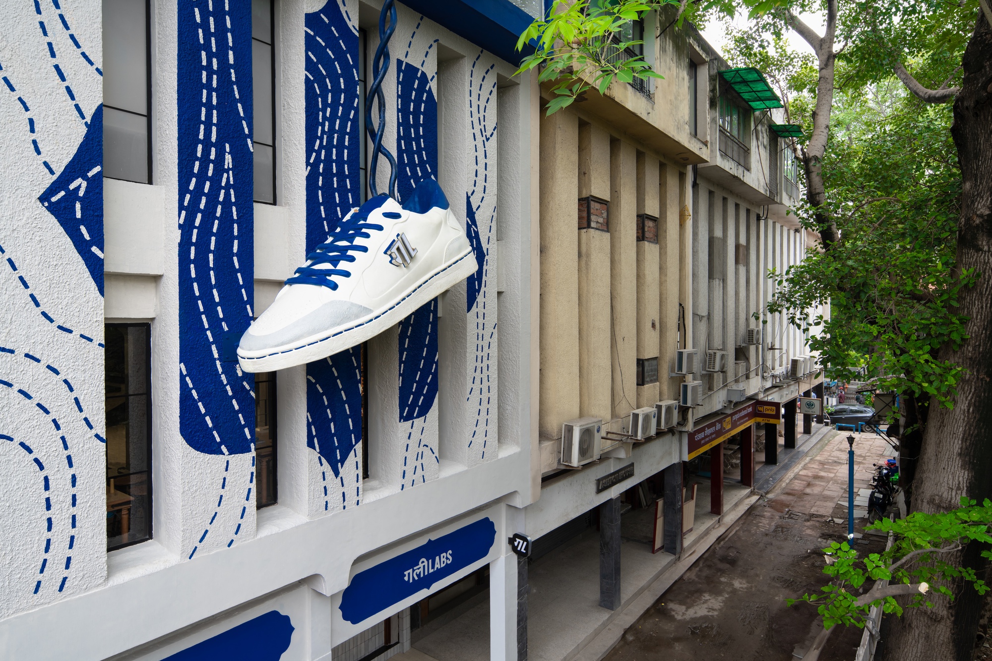 Angled street view showing the mural facade with oversized sneaker installation beneath overhanging tree branches