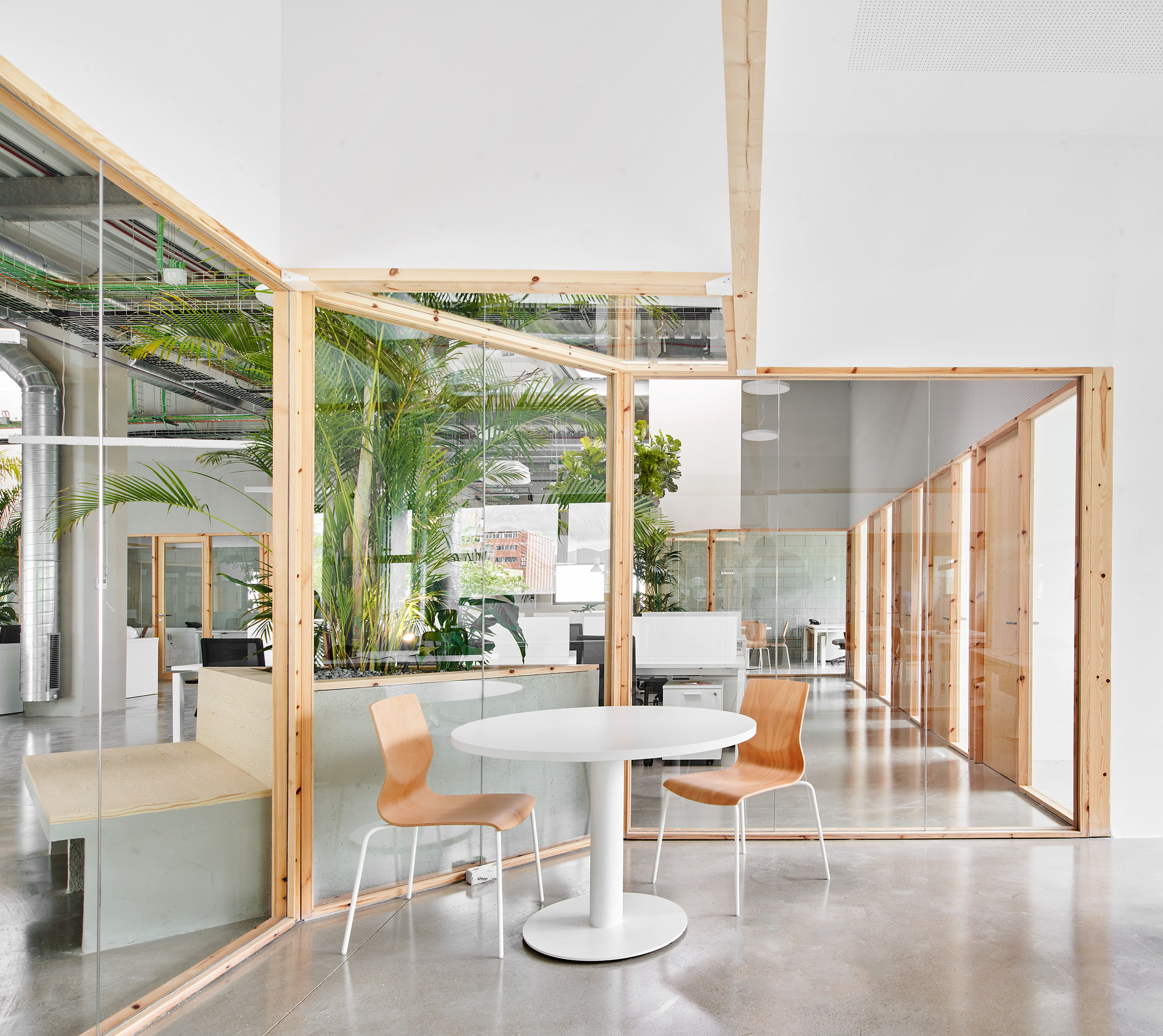 Glass-enclosed timber-framed meeting room with potted palms and exposed mechanical ducts overhead