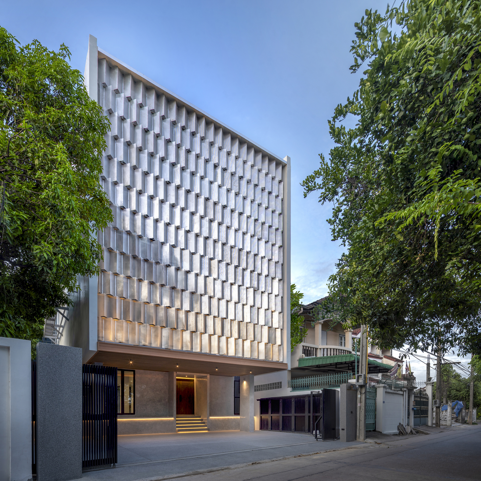 Facade at dusk showing the illuminated entrance and vertical brise-soleil screen framed by trees