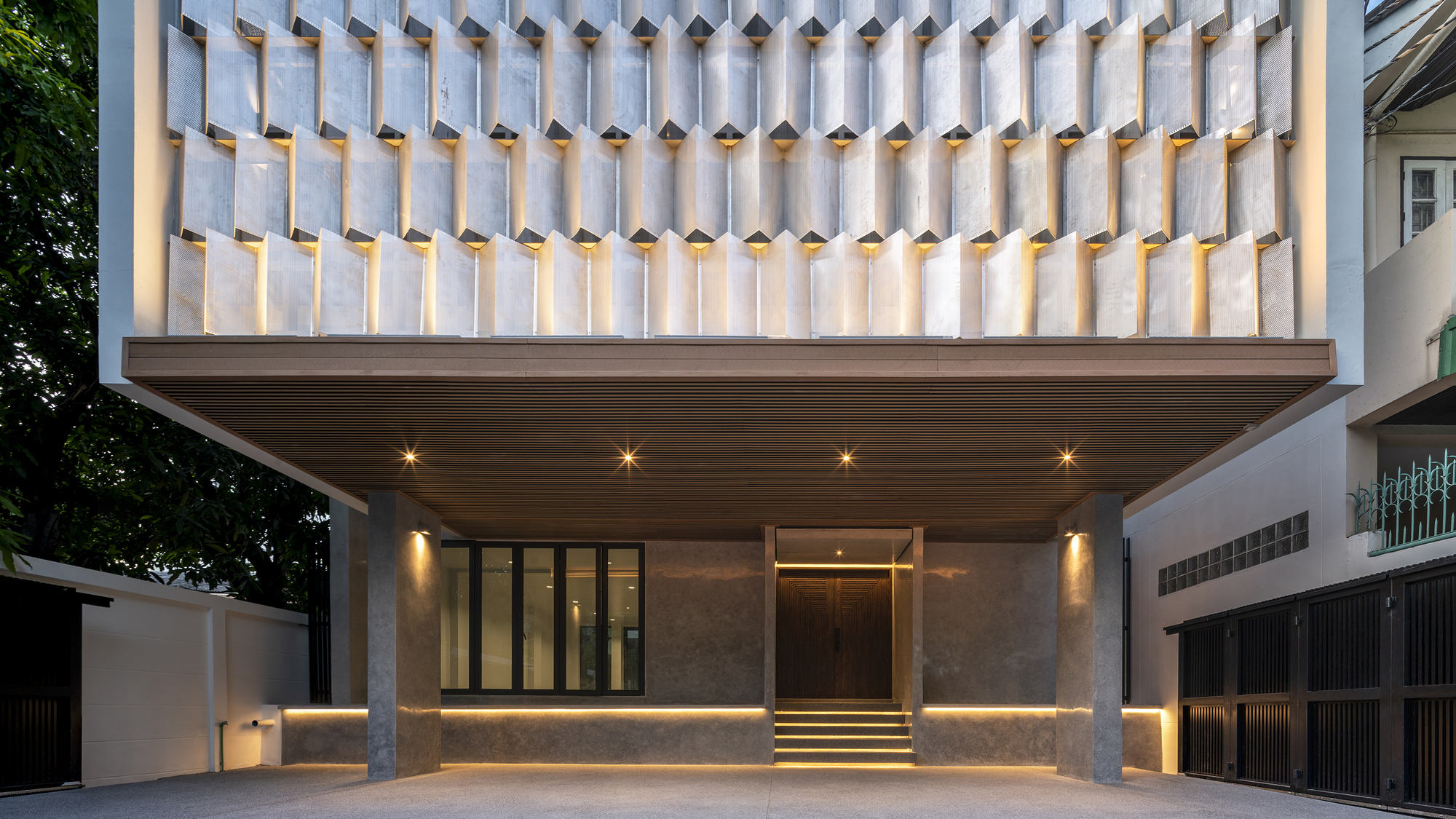 Street facade with cantilevered upper volume clad in concrete fins above an illuminated entry at twilight