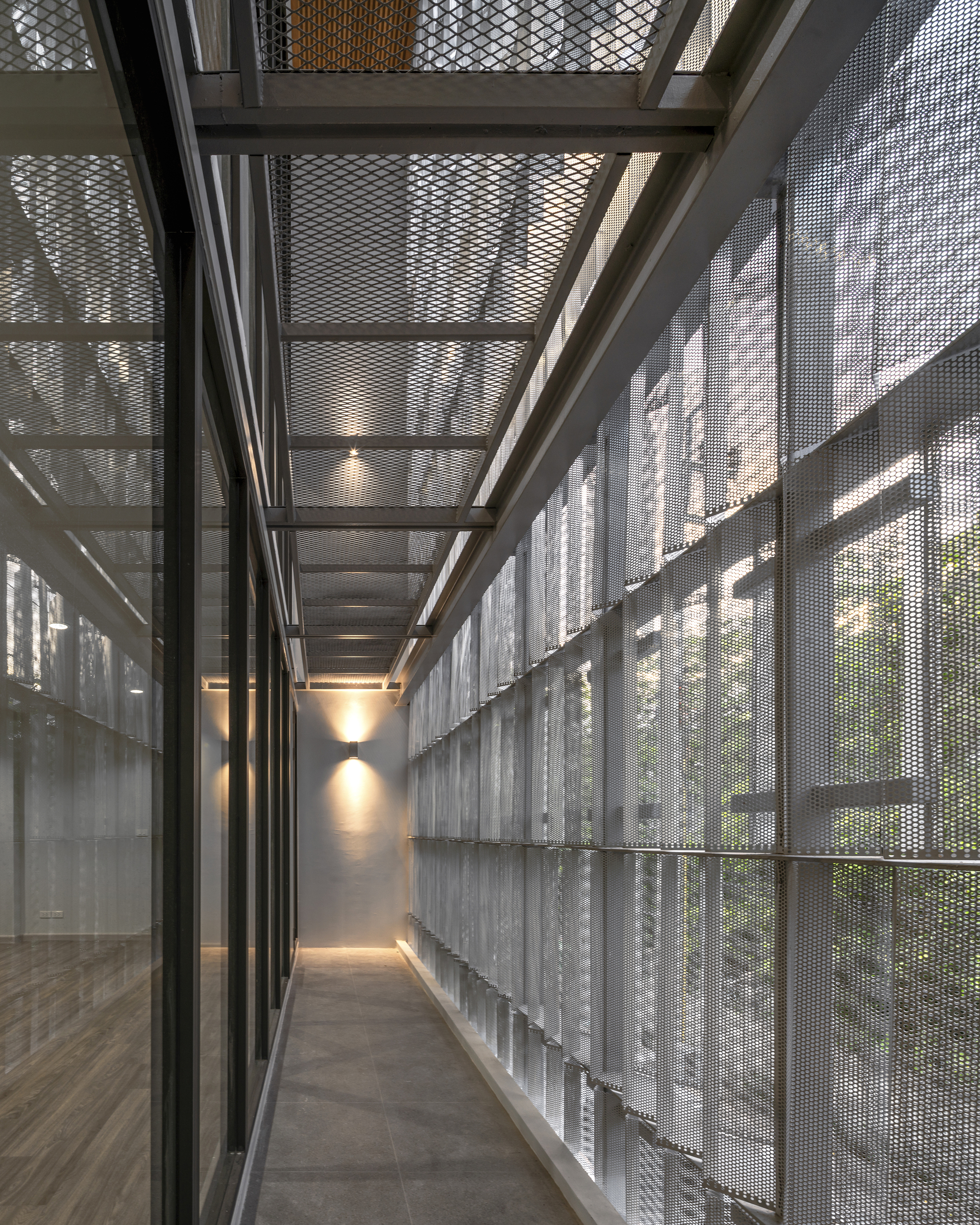 Corridor enclosed by perforated metal screens and exposed steel beams filtering dappled light from exterior planting
