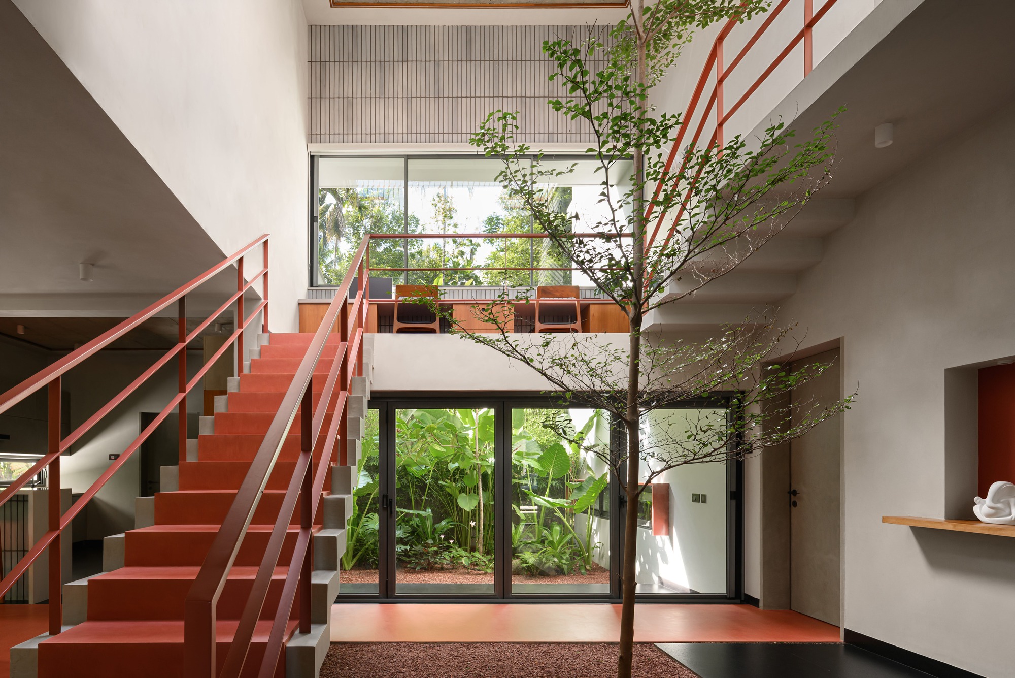 Red metal staircase and interior tree framed by concrete beams and glazed garden views