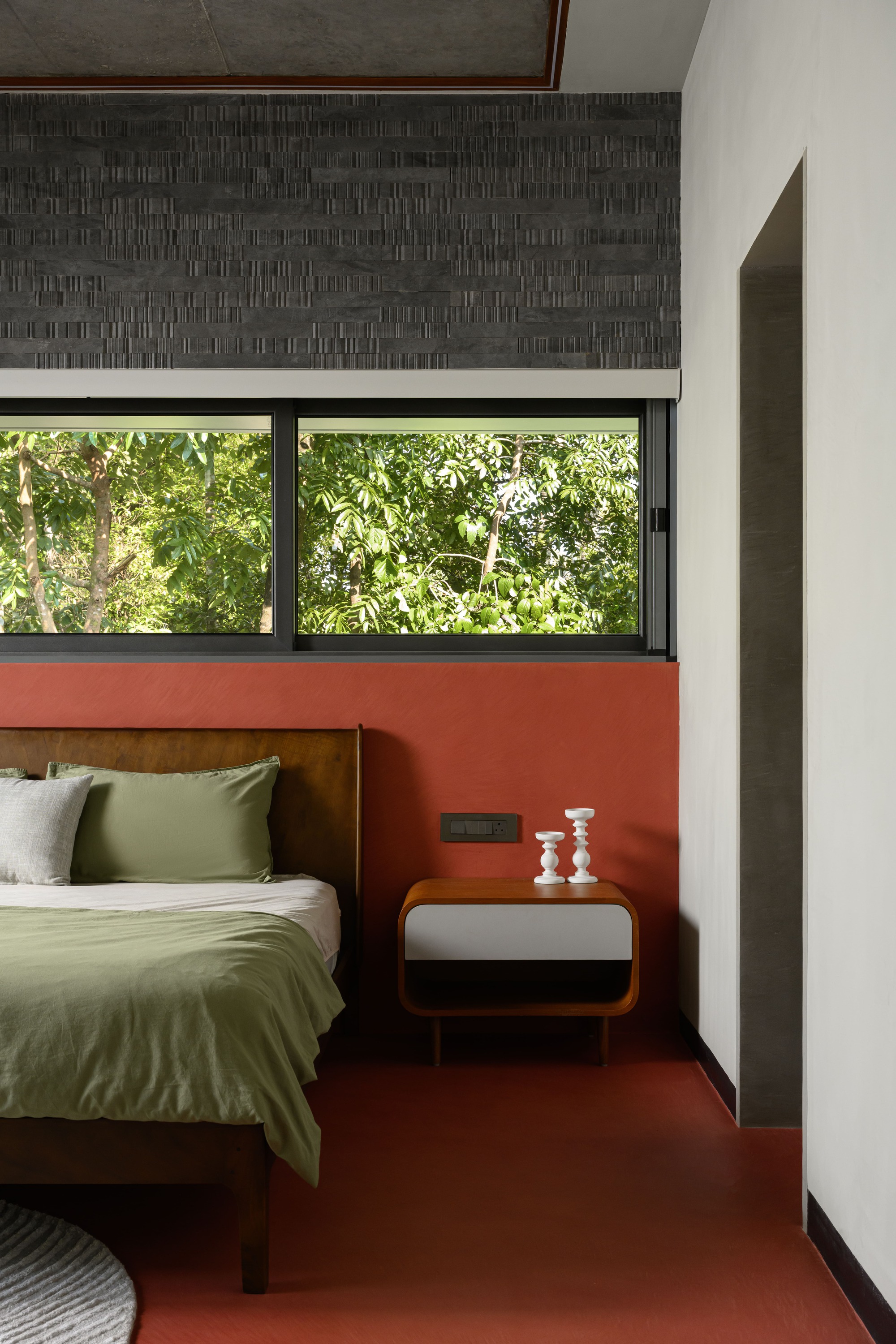 Bedroom with terracotta floor and horizontal window overlooking dense foliage framed by textured dark tile band