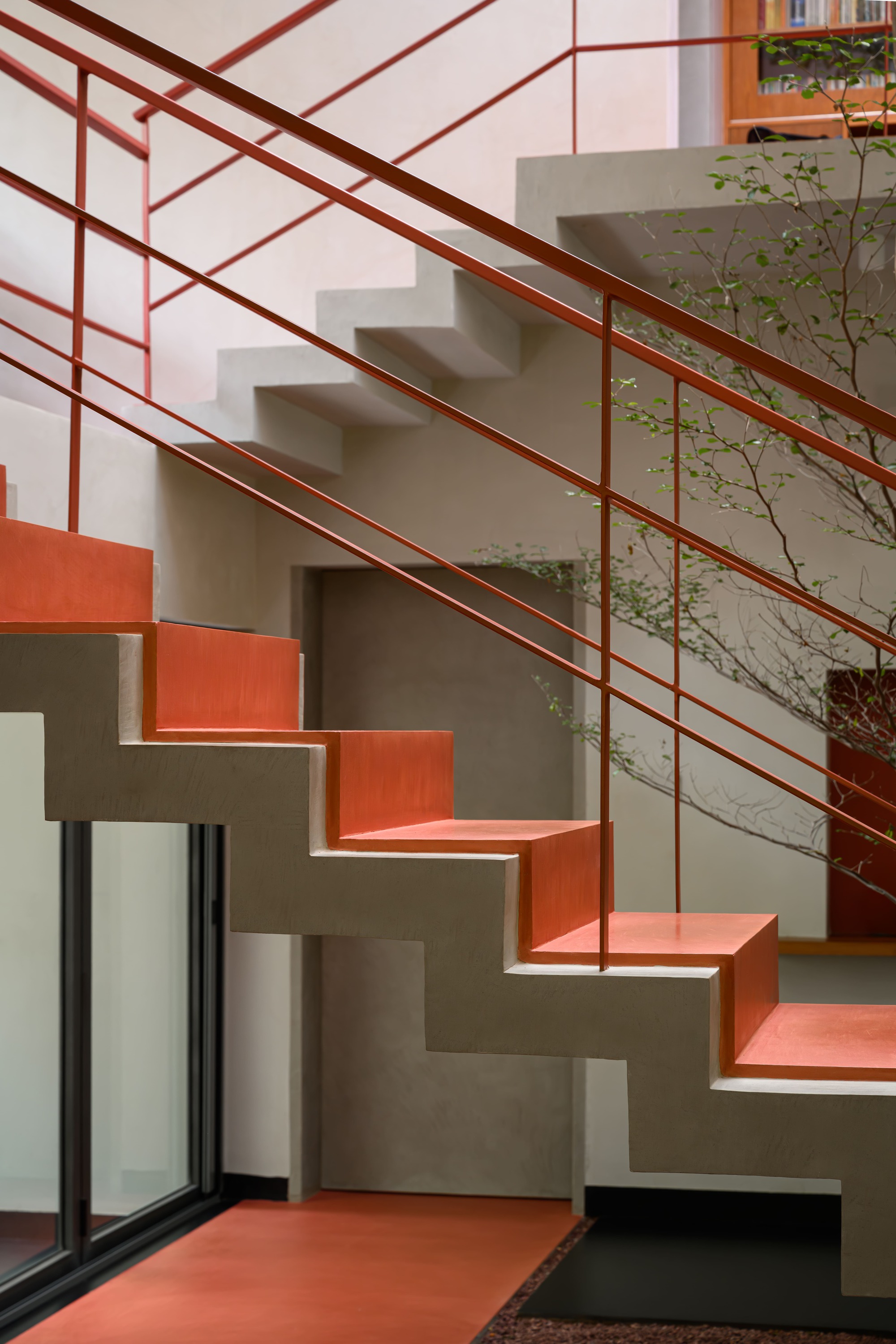 Cantilevered concrete staircase with coral-painted treads and red tubular steel railing beside a planted interior courtyard