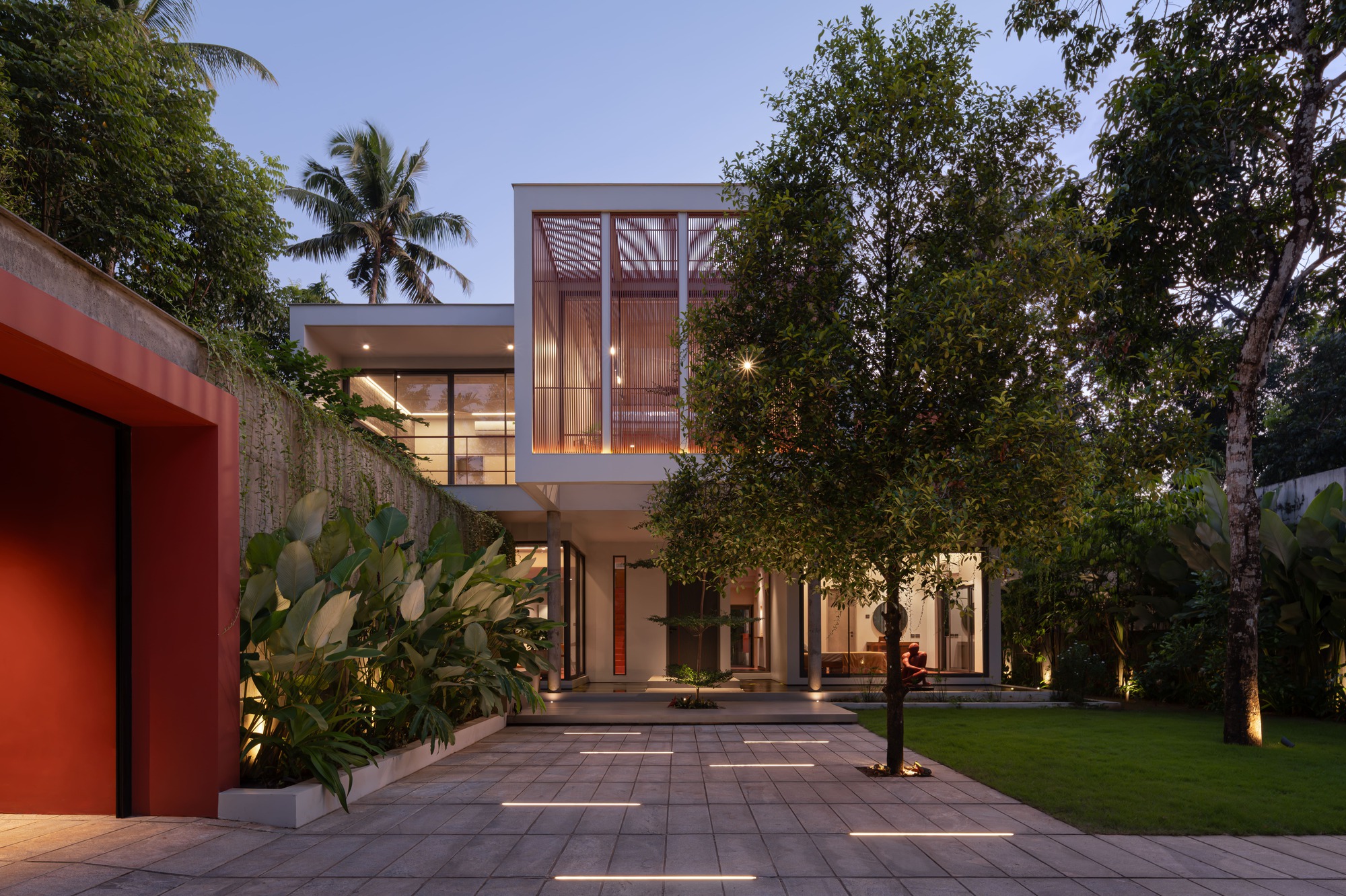Front courtyard view at dusk showing the two-storey facade with timber screens and uplighting among mature trees