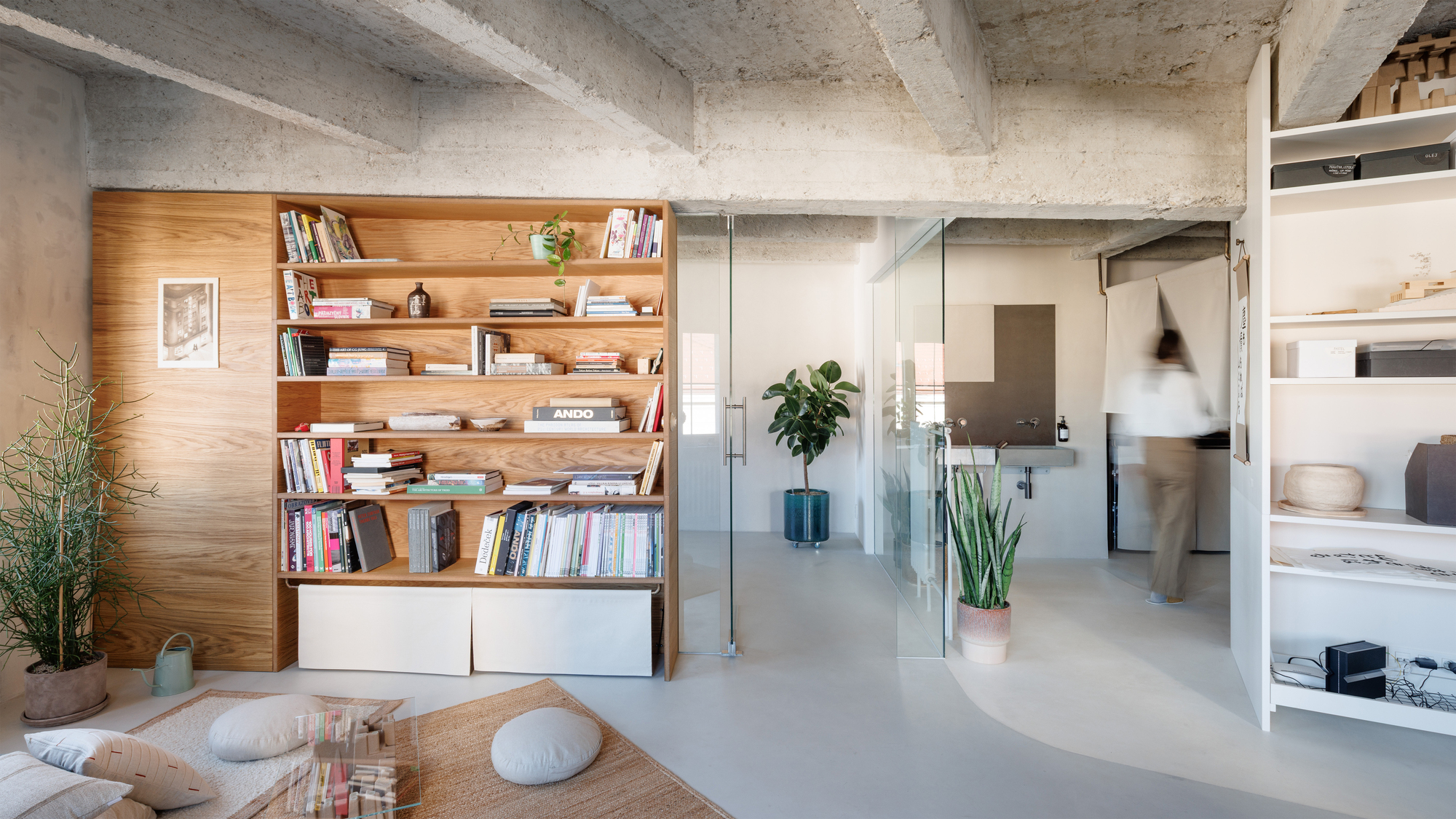 Timber bookshelf wall beside a glass partition with a figure walking through the background