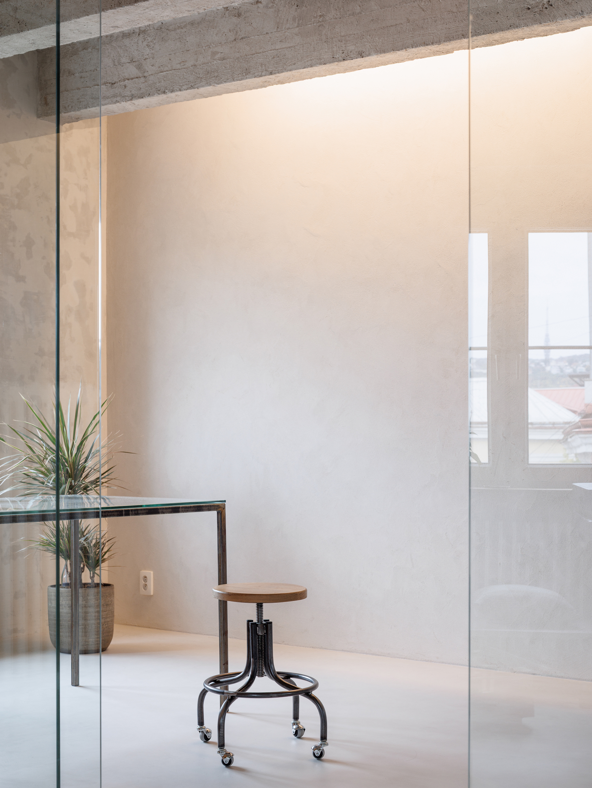 Interior workspace with glass partition, adjustable stool, potted plant, and exposed concrete ceiling beams