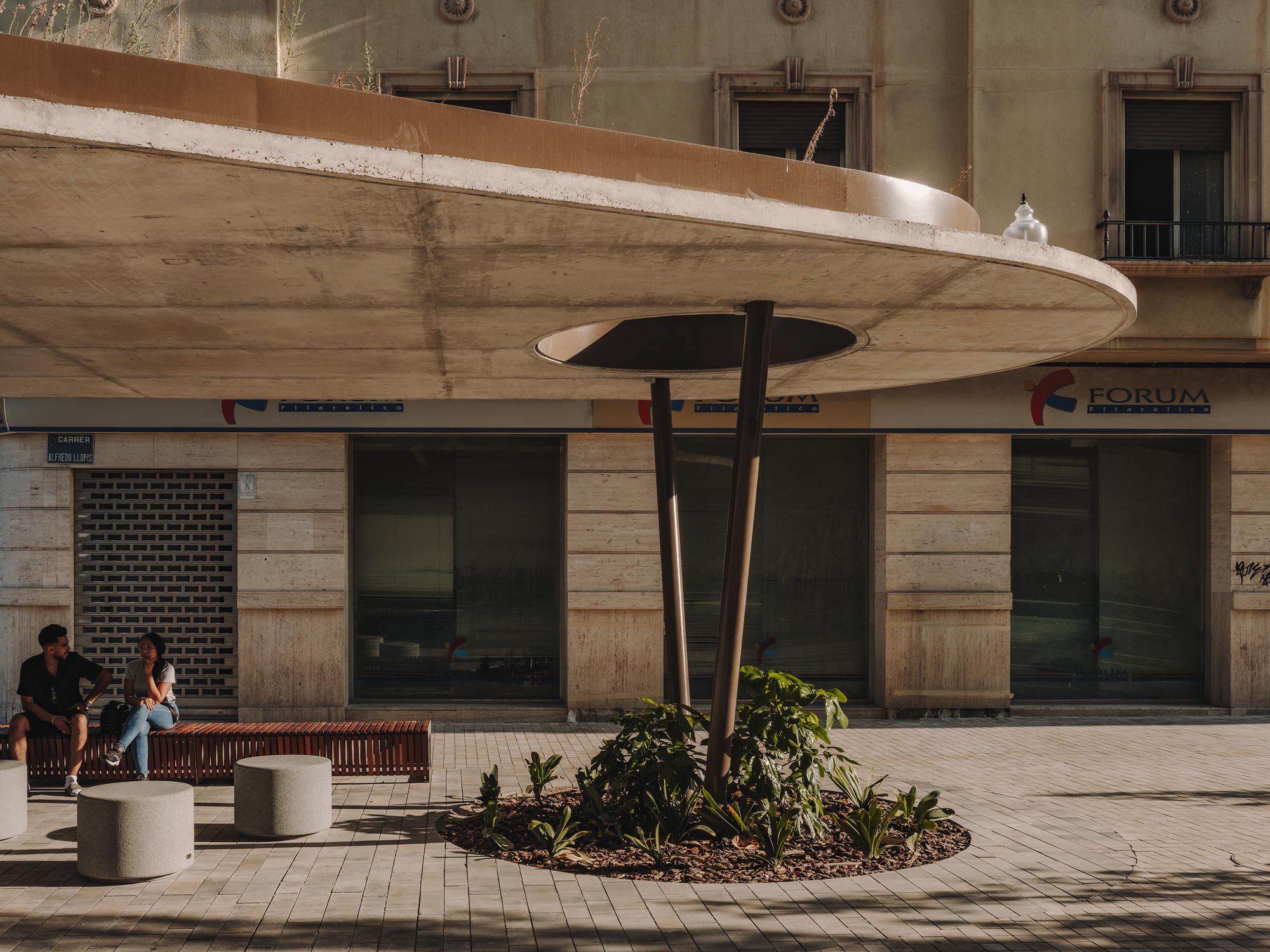 Concrete canopy with circular opening and central support column beside timber bench seating at dusk