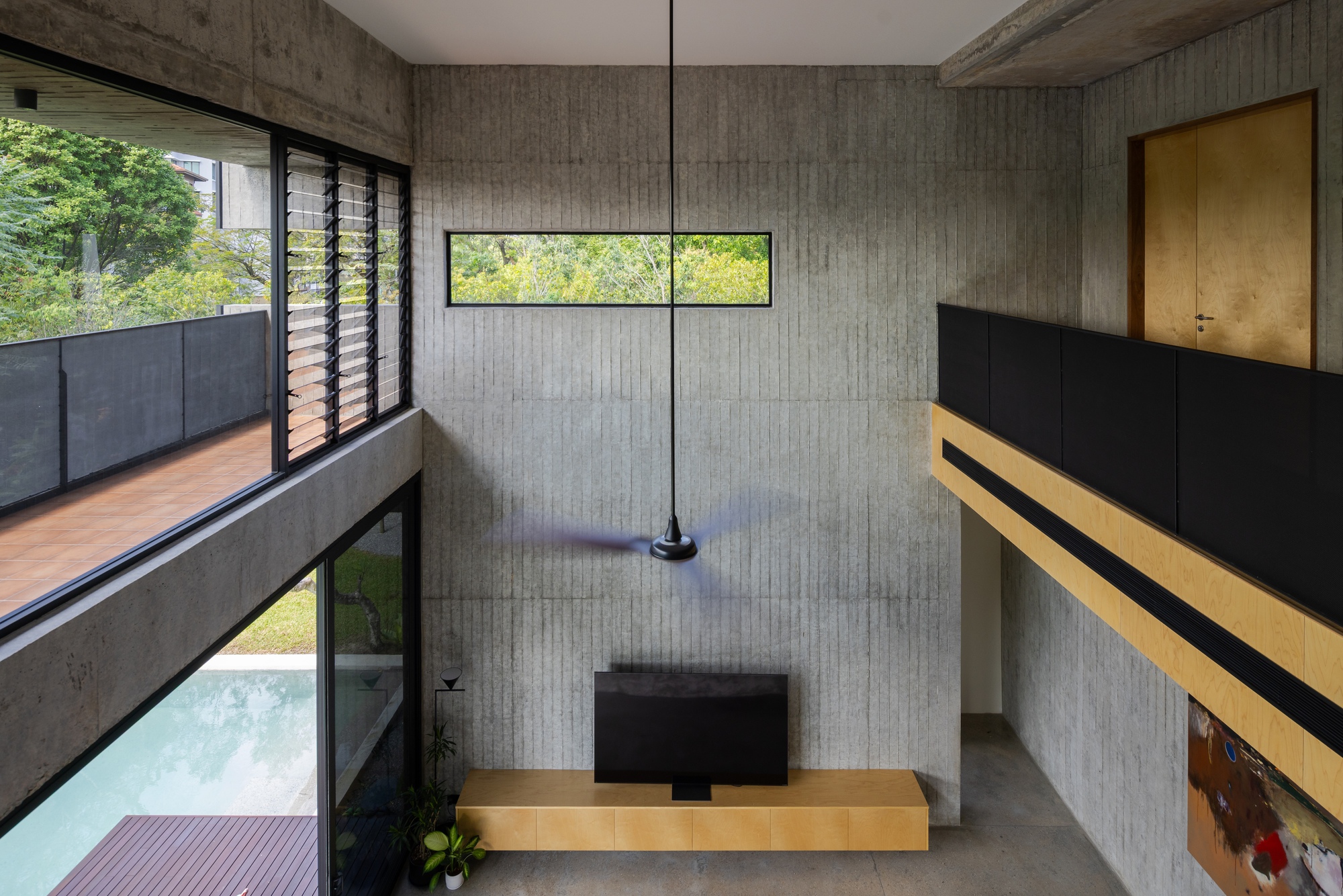Interior void with board-formed concrete walls, horizontal ribbon window, pendant light, and timber bridge spanning overhead