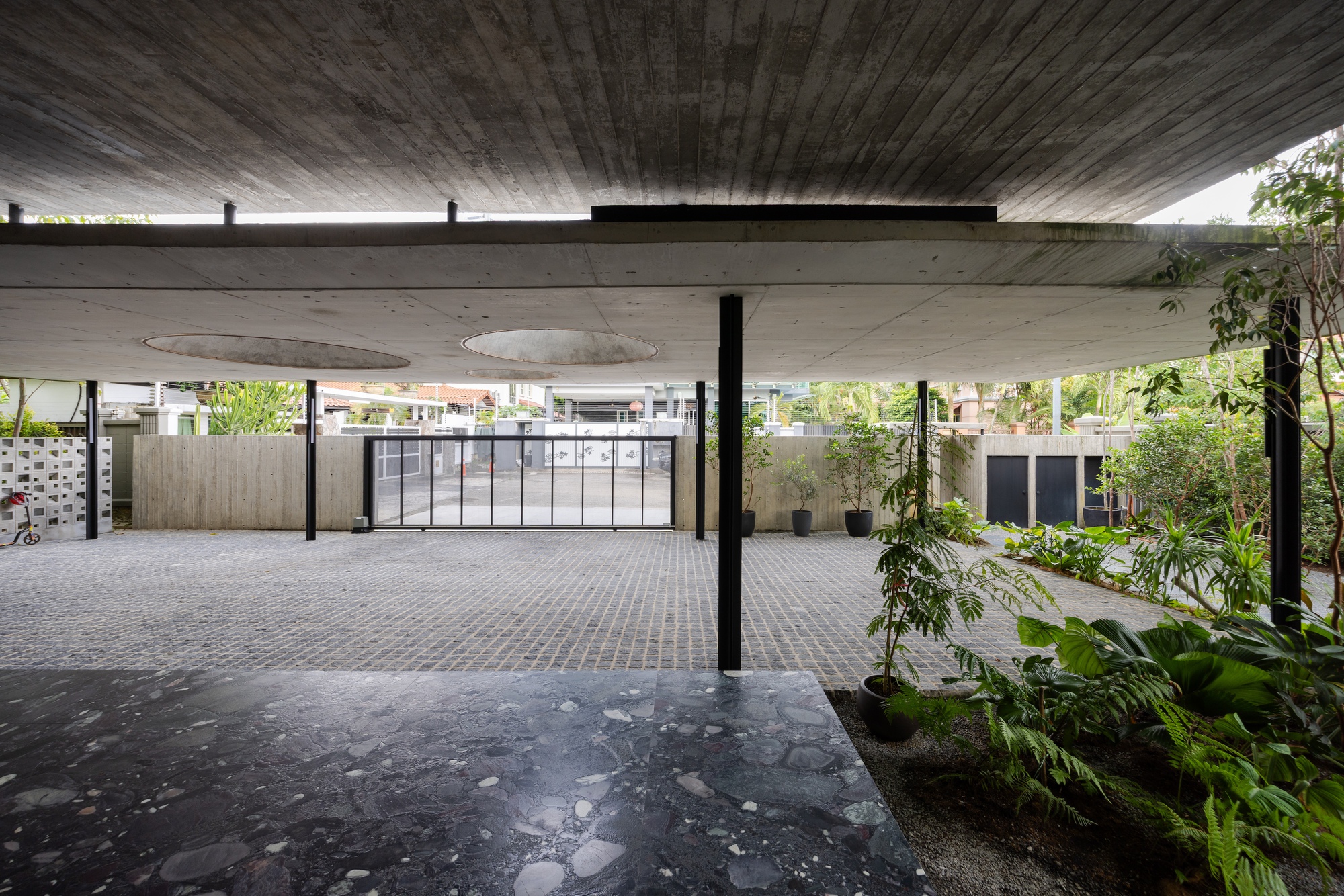 Covered carport with exposed concrete ceiling and circular skylights looking toward planted beds and storage units