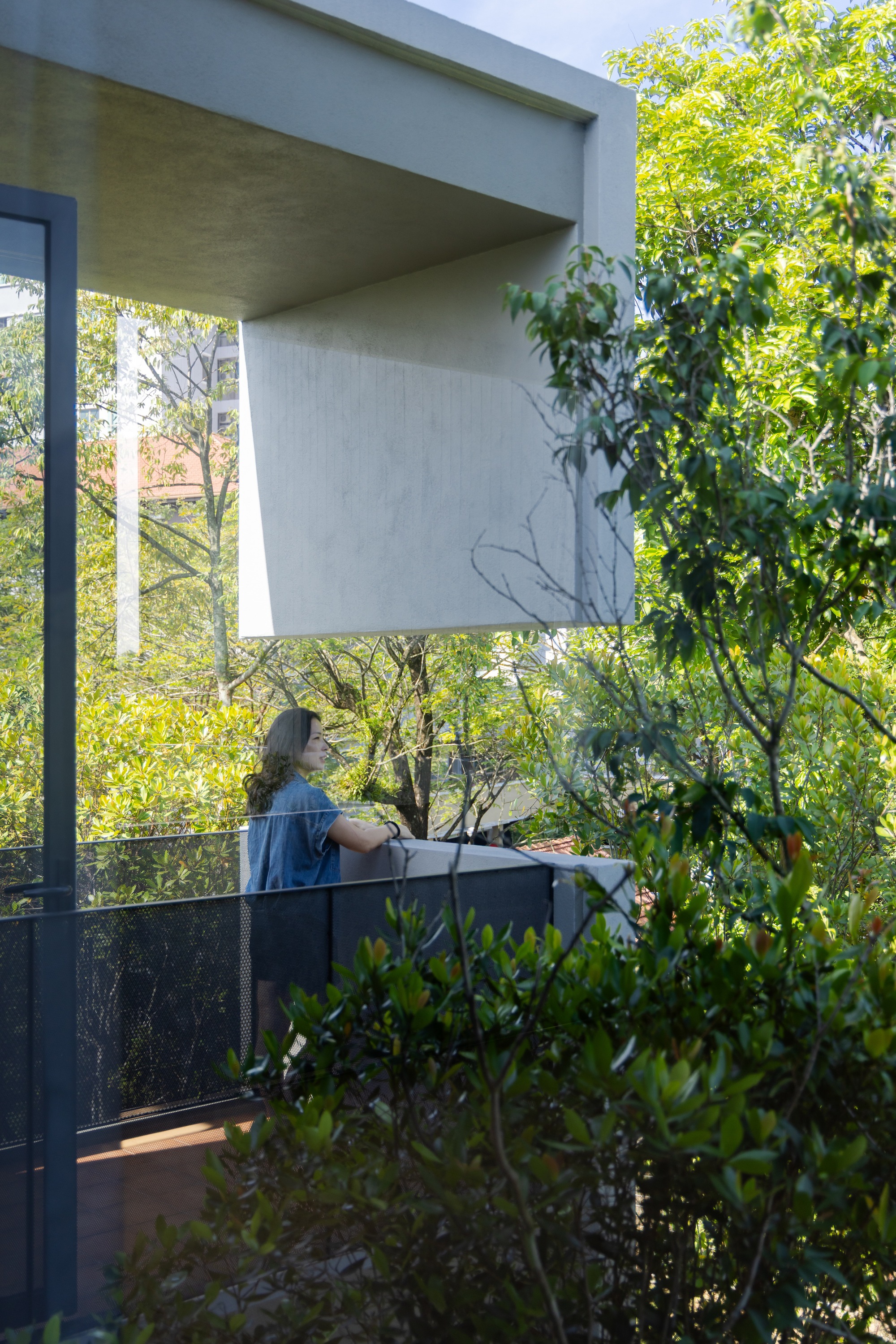 Covered balcony with person tending plants behind a dark metal railing amid dense foliage