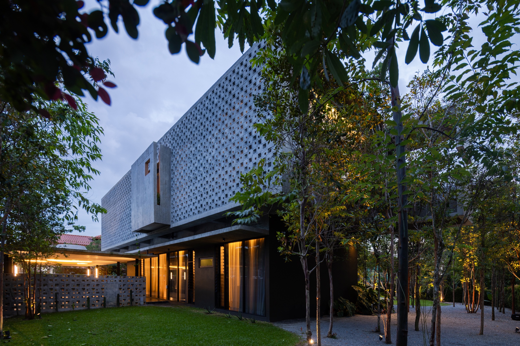 Perforated white concrete volume above glass-walled base surrounded by illuminated trees at dusk