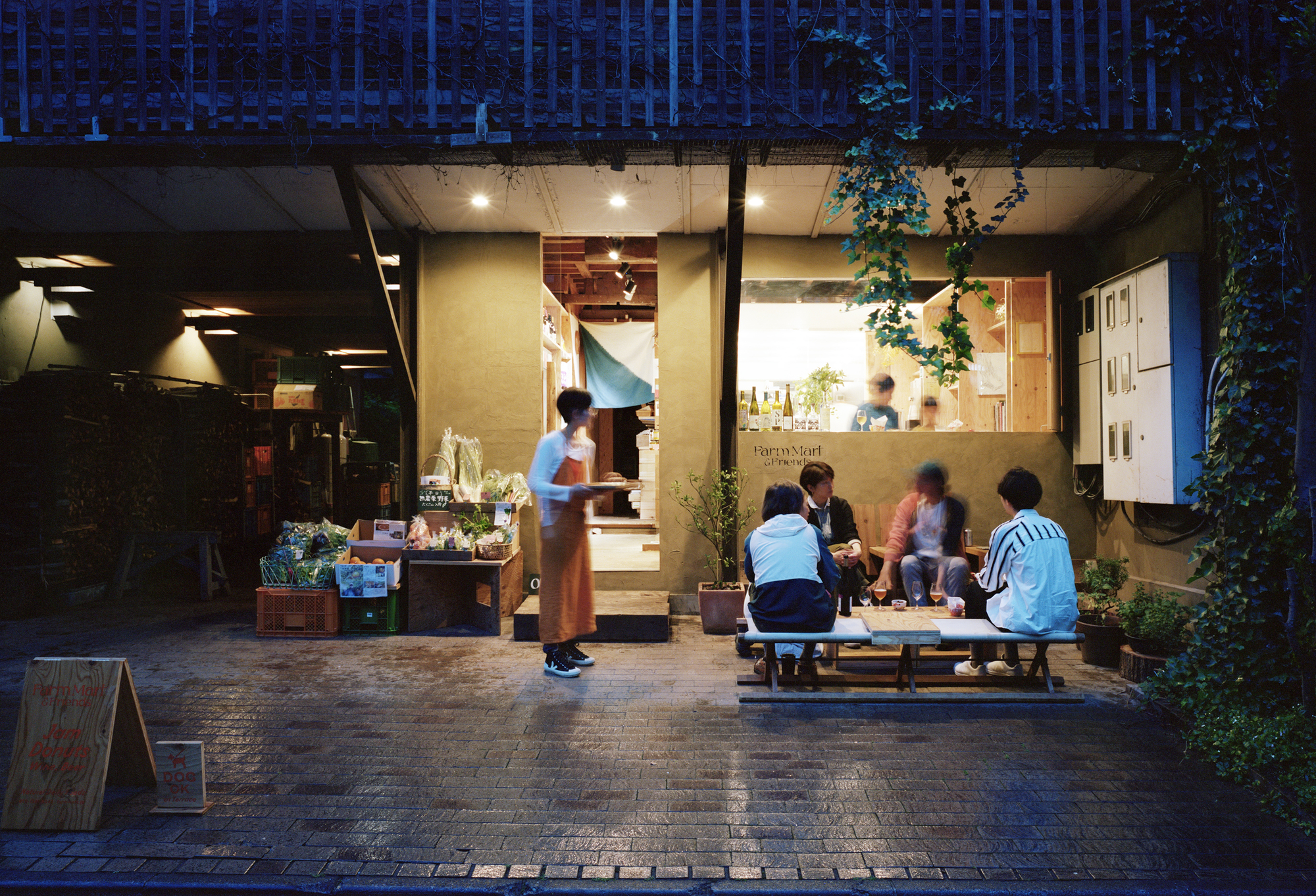 Glass-fronted shopfront opening to a paved courtyard with people gathered at dusk