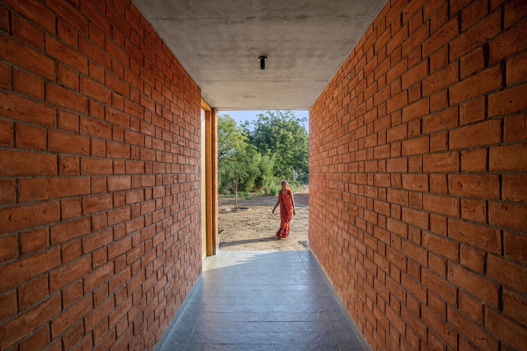 Corridor framed by exposed red brick walls leading to a figure in an orange sari