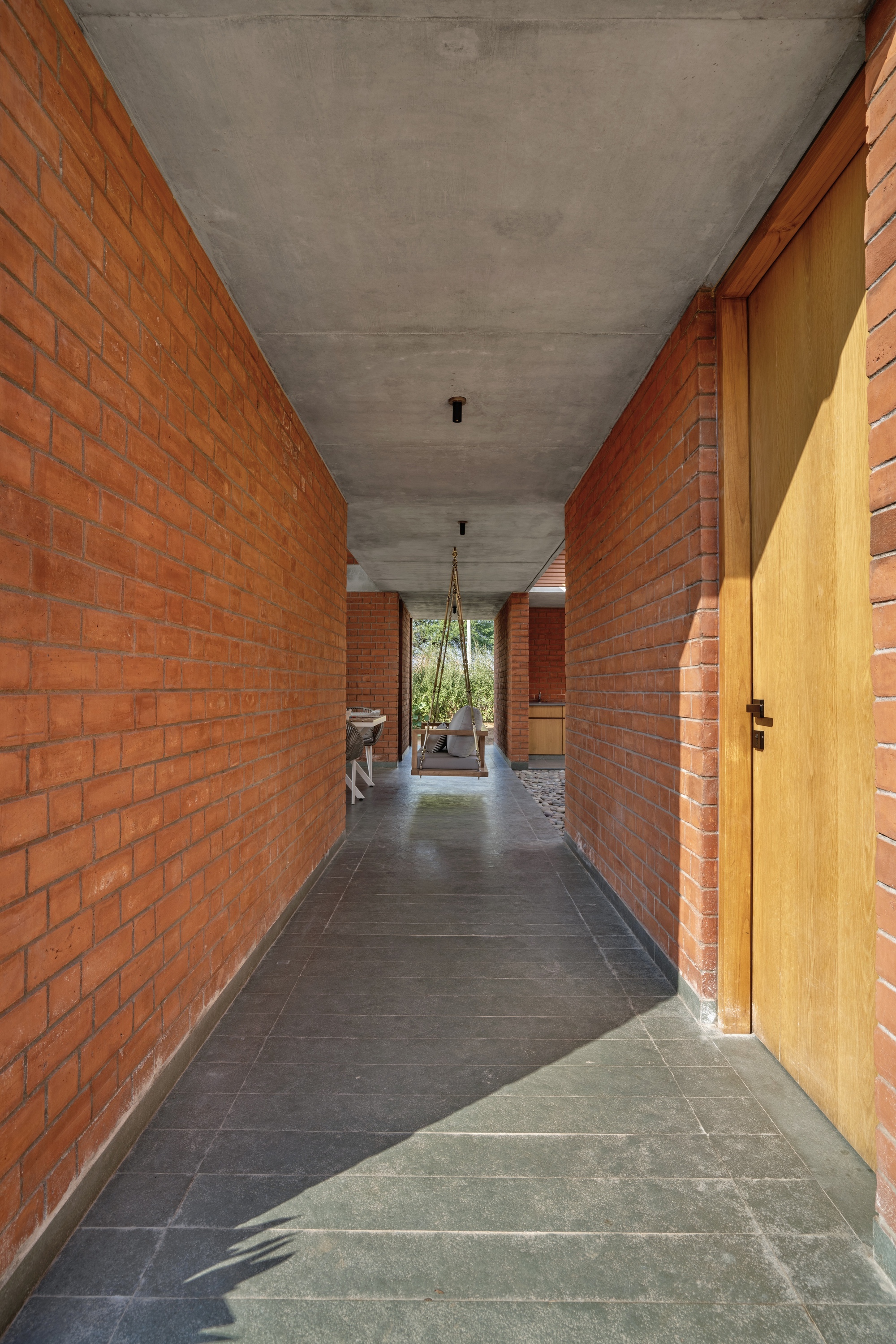 Covered corridor with red brick walls, concrete ceiling, and slate flooring leading to a planted courtyard