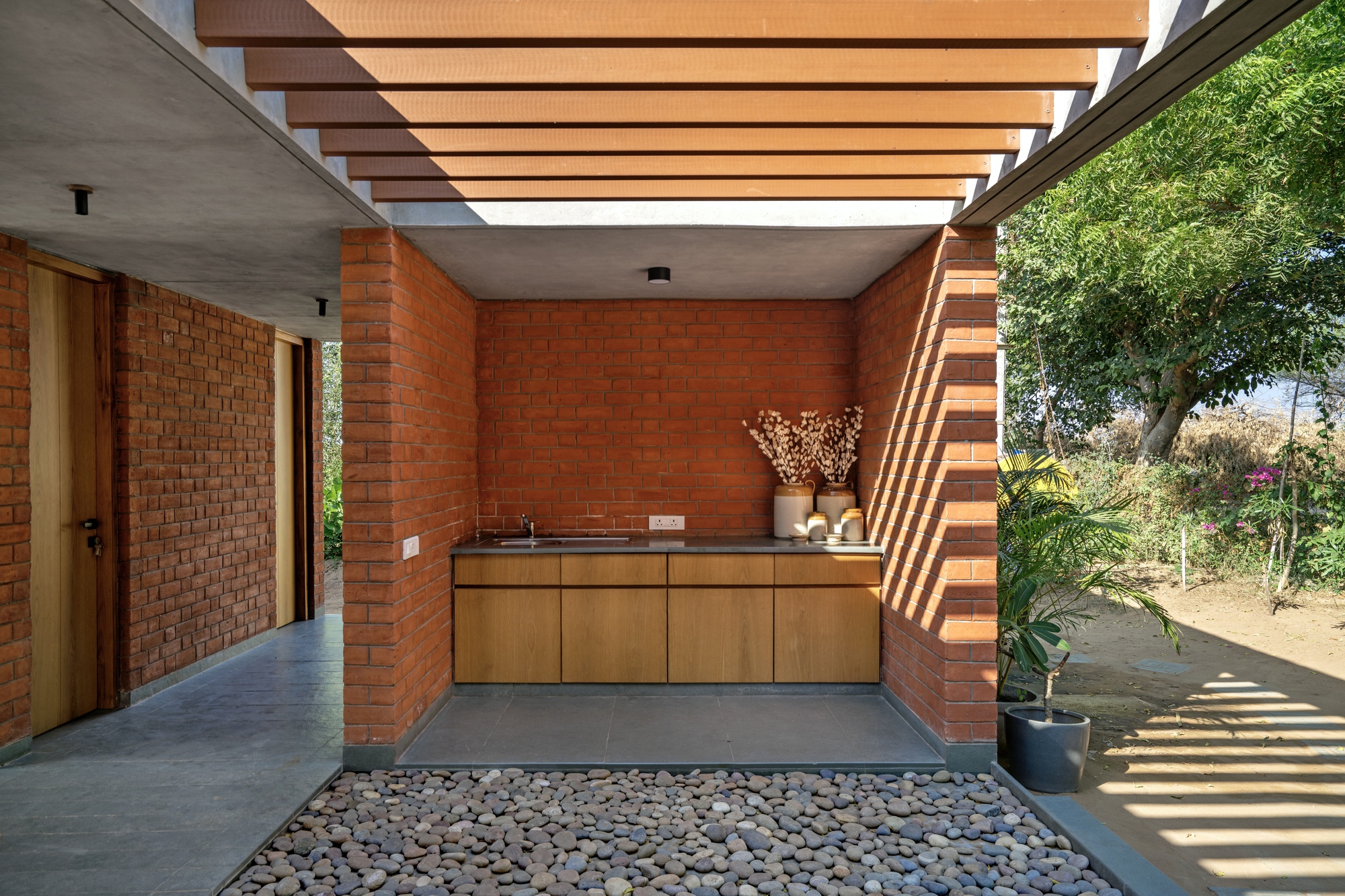 Semi-outdoor kitchen counter beneath timber pergola slats with river stone flooring and afternoon shadows