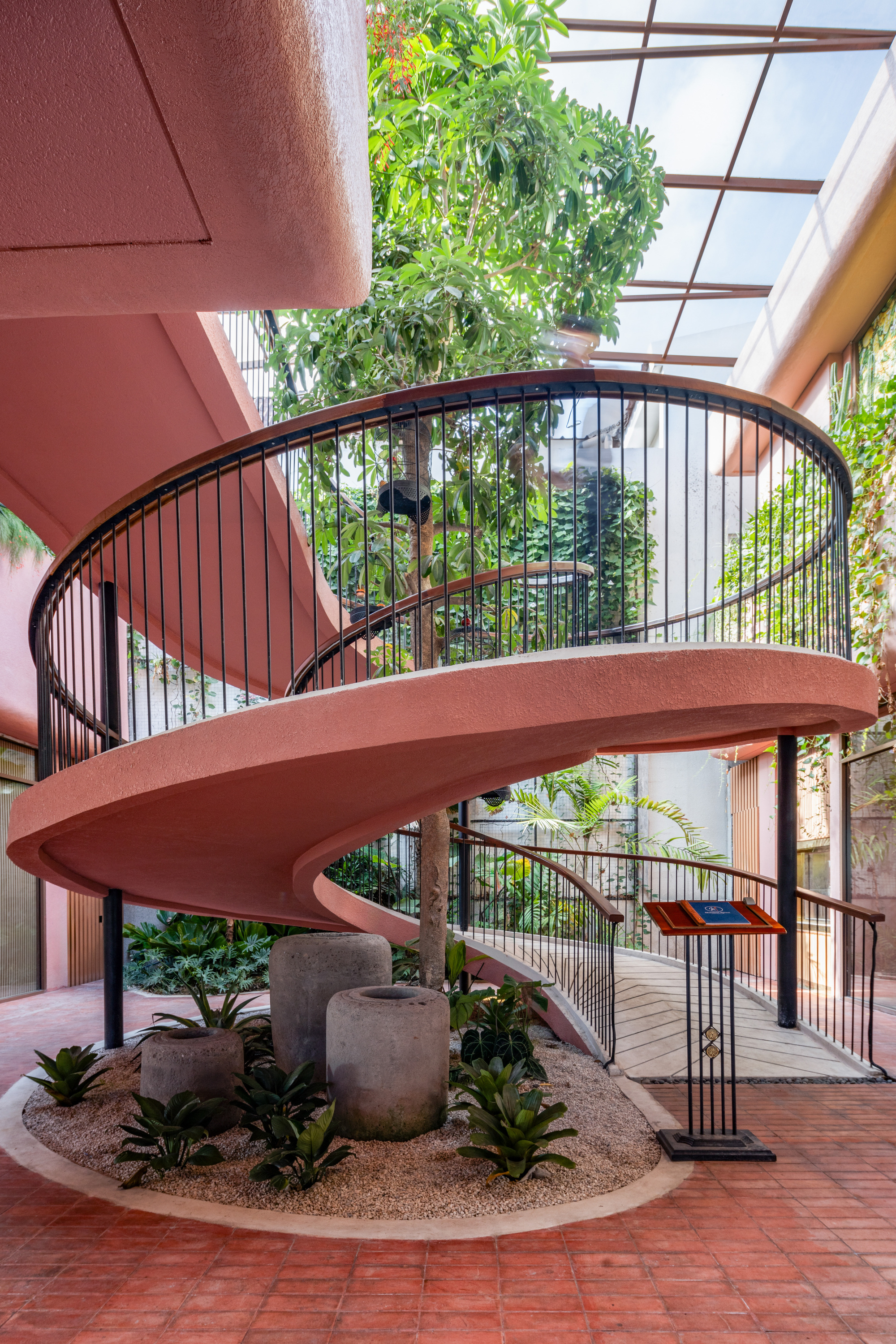 Spiral staircase with black metal railings and pink concrete treads beneath a glass roof with hanging vines