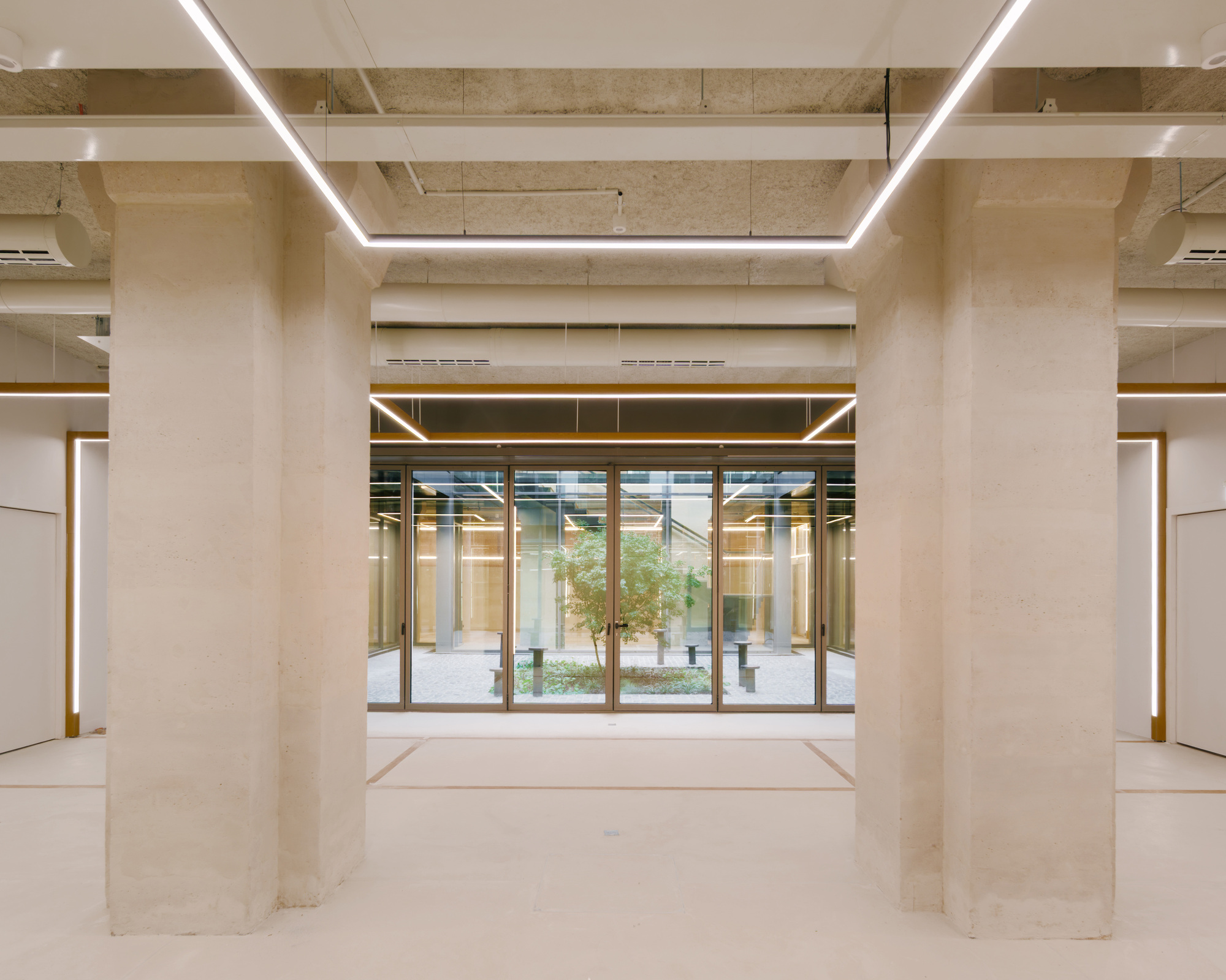 Interior space with exposed concrete ceiling beams and rectangular light fixtures framing a glazed courtyard entrance