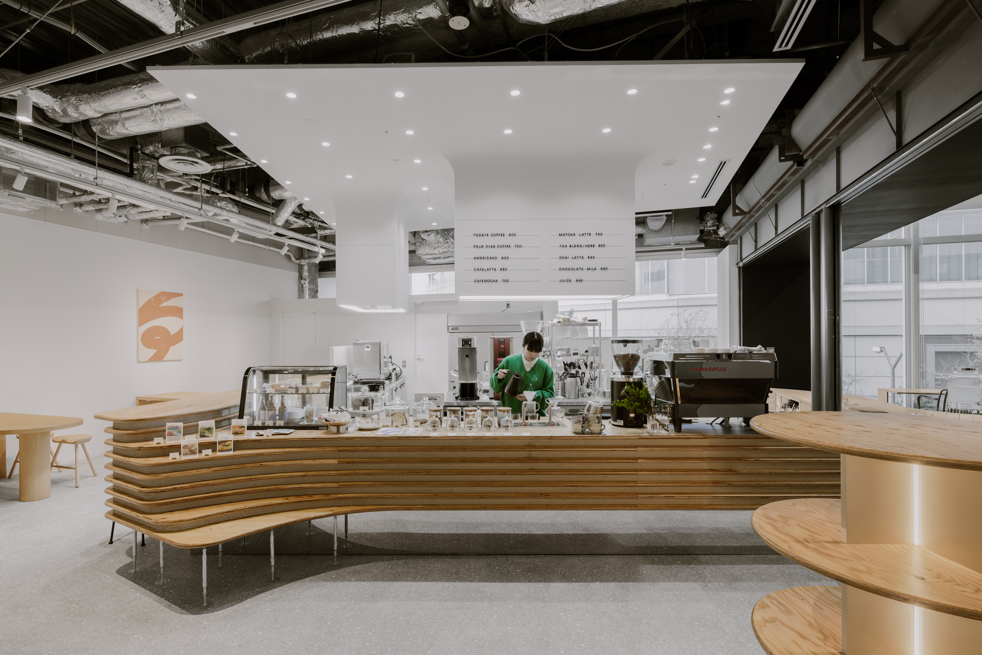 Cafe counter with layered plywood front and baristas working under exposed mechanical services and white menu boards