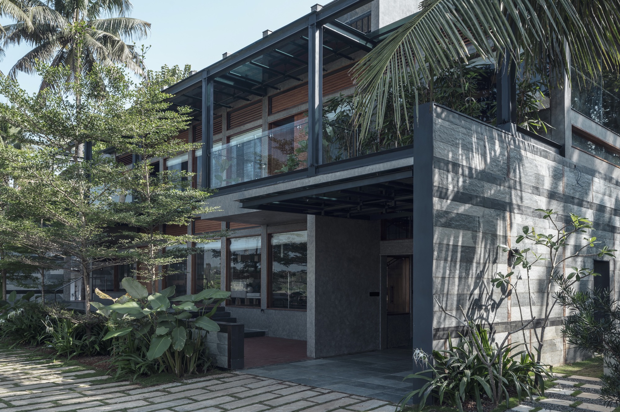 Two-story concrete and timber facade with steel frame pergola surrounded by palm fronds and tropical planting