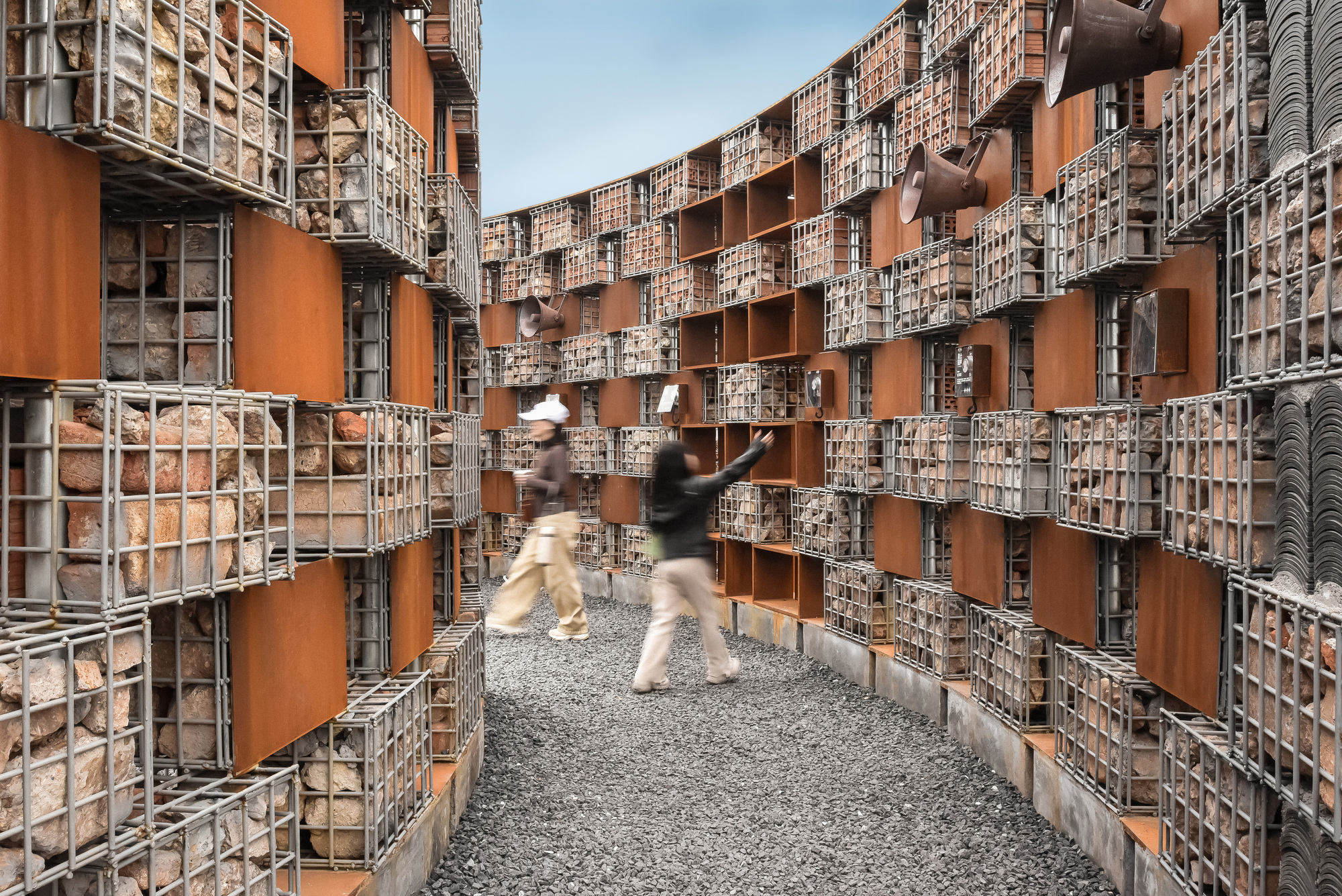 Narrow gravel corridor flanked by gabion walls filled with stone and terracotta panels as two visitors walk through