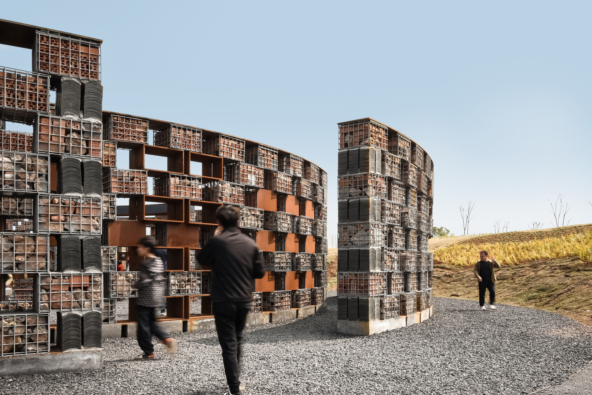 Curved gabion walls with stone fill and terracotta openings on a hillside with dry grass and bare trees