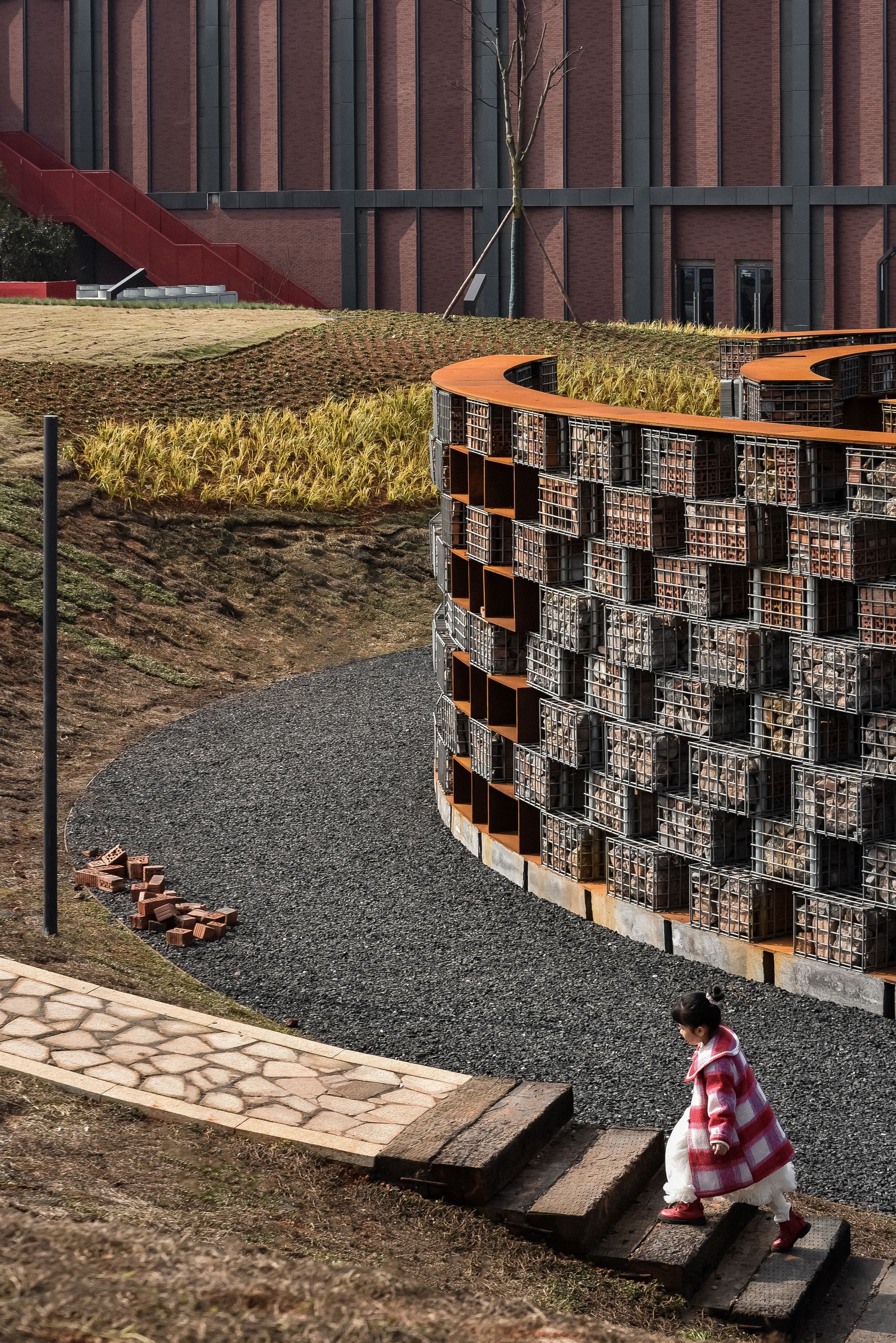 Visitor in a red jacket descending stone steps beside a curving gabion wall with golden grasses above