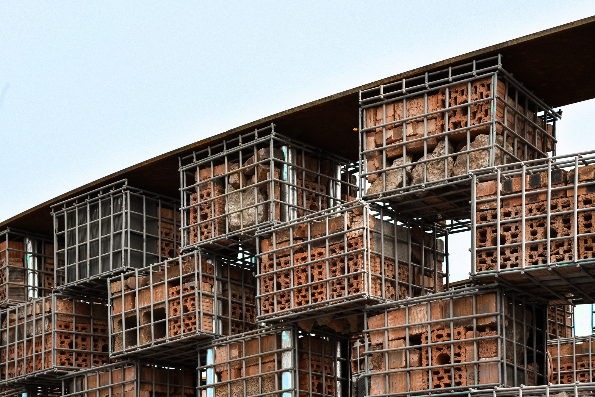 Stacked gabion crate facade with stone fill and steel mesh beneath a cantilevered corten roof