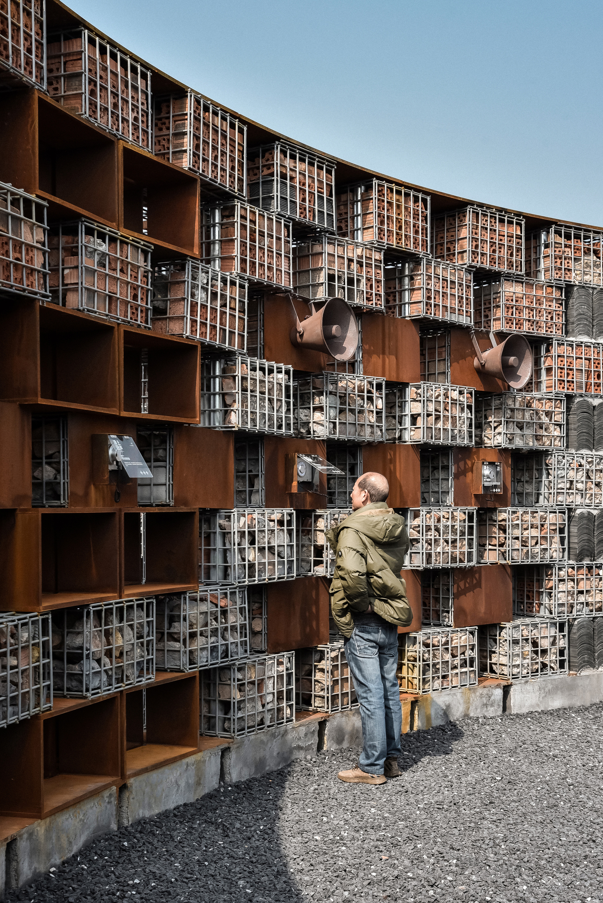 Visitor photographing the curved facade composed of wire-filled gabion baskets, corten frames and mounted speaker horns