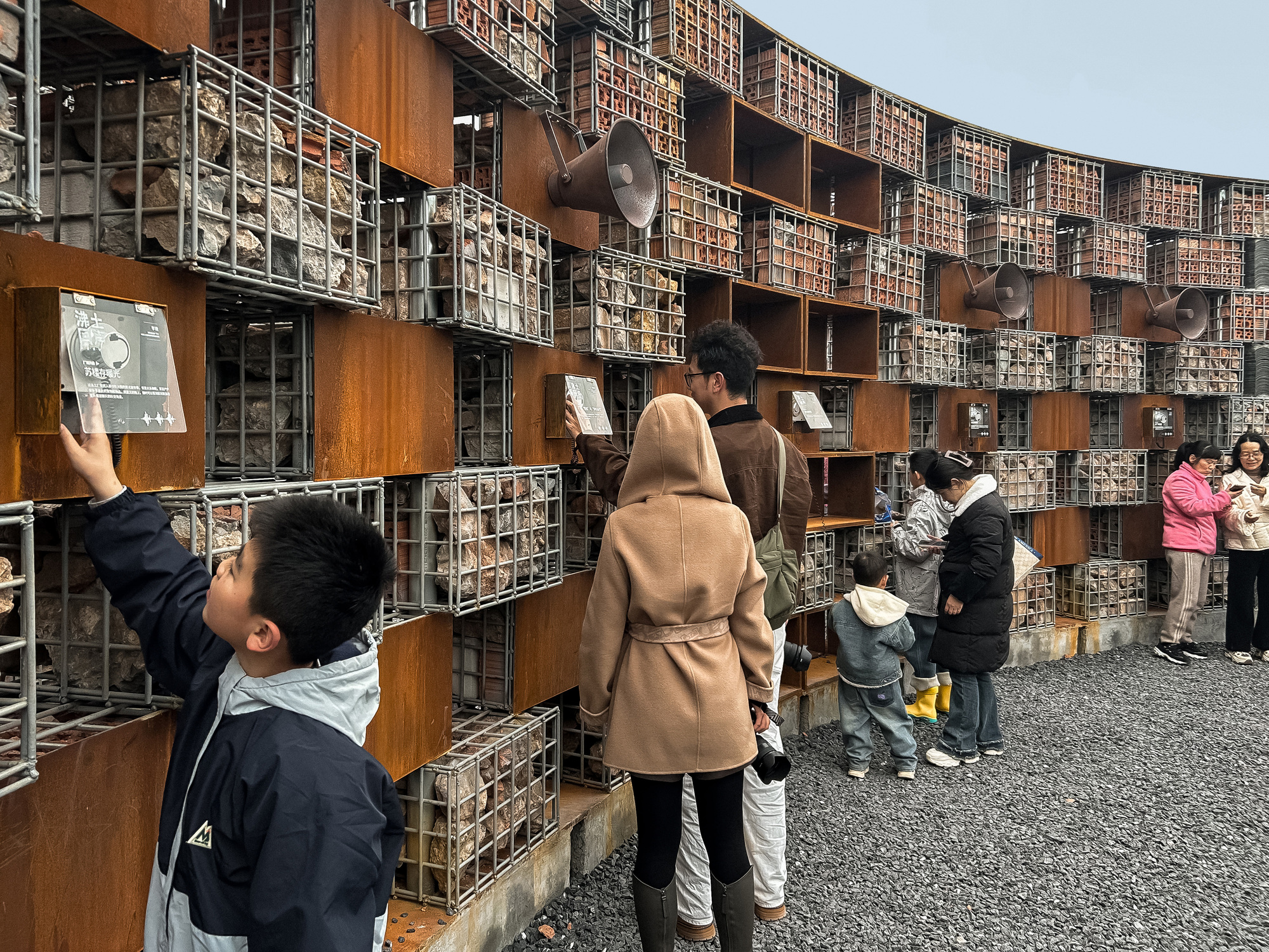 Families examining display niches set into the gabion and corten steel grid wall along the gravel plaza