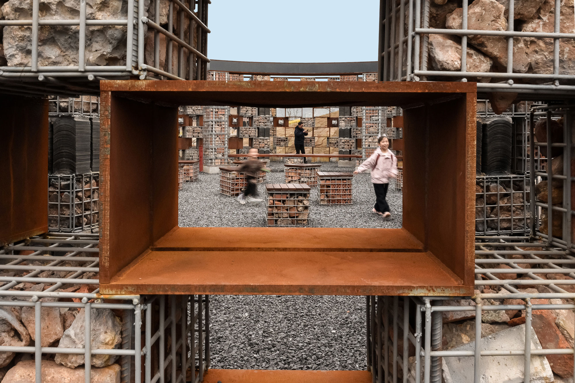 View through a weathered steel frame opening showing visitors in the courtyard beyond