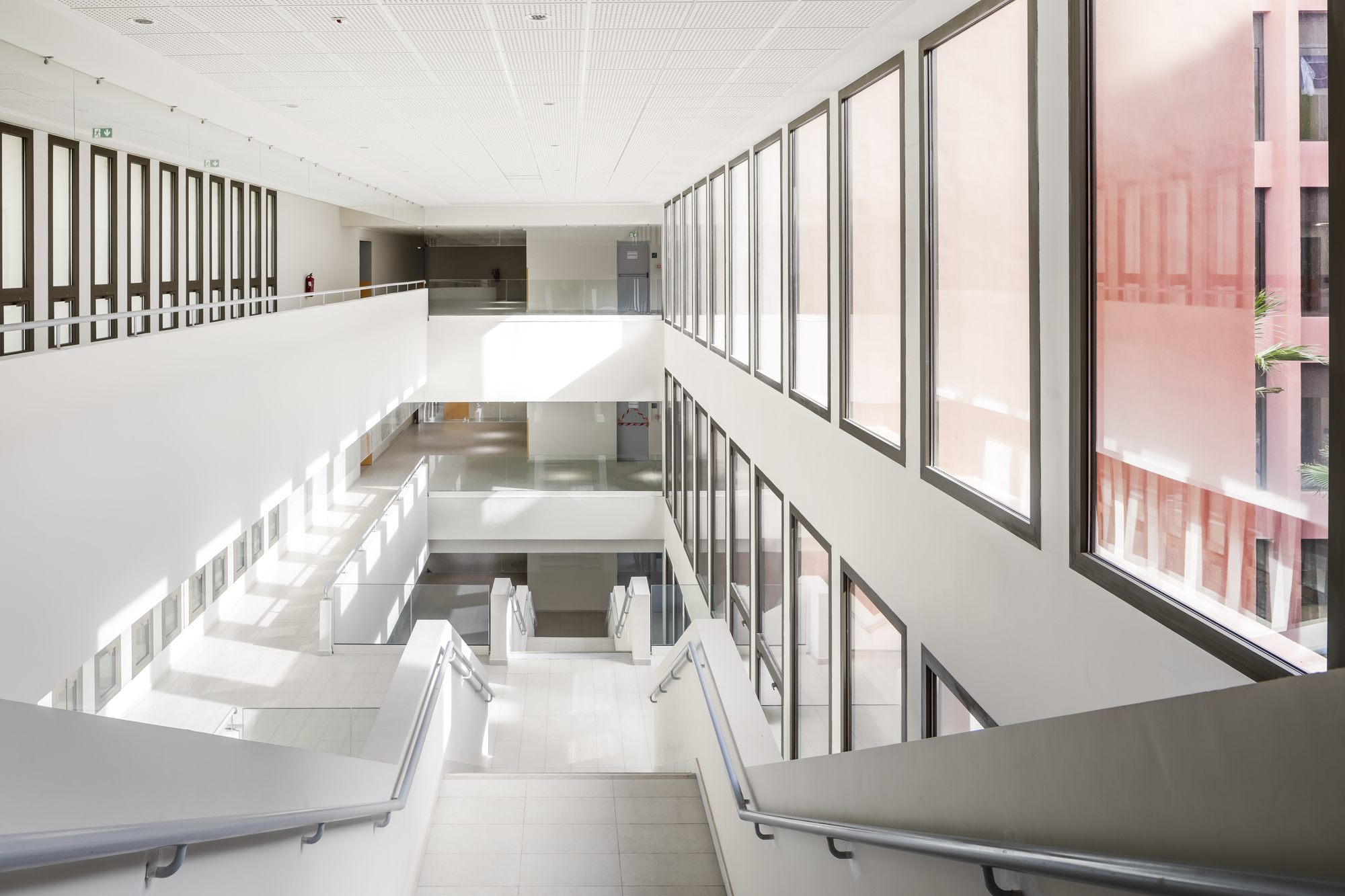 Interior atrium with white walls, continuous glazing overlooking a courtyard, and a staircase descending three levels