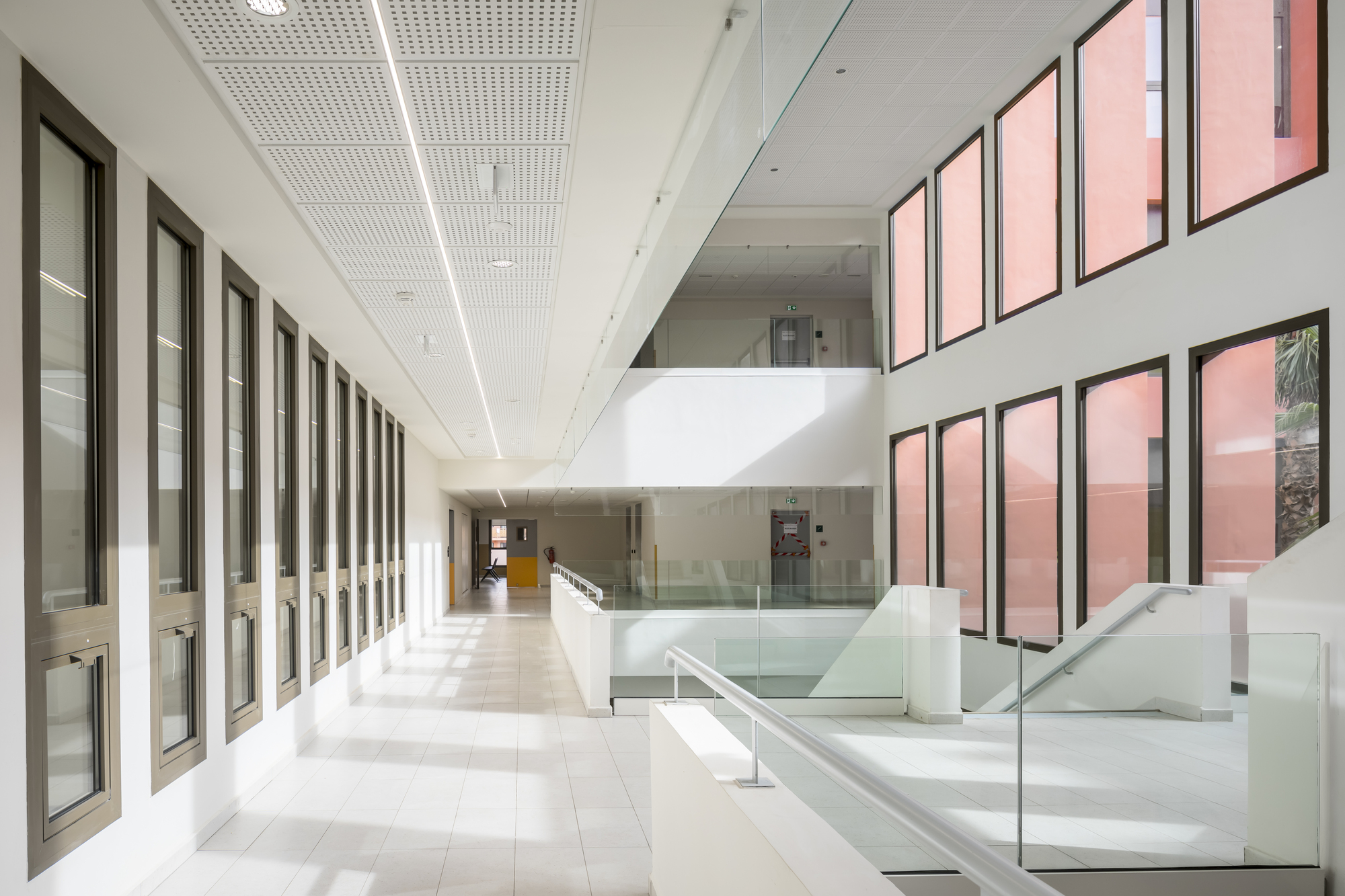 White tiled corridor with repetitive window frames and perforated ceiling panels under daylight
