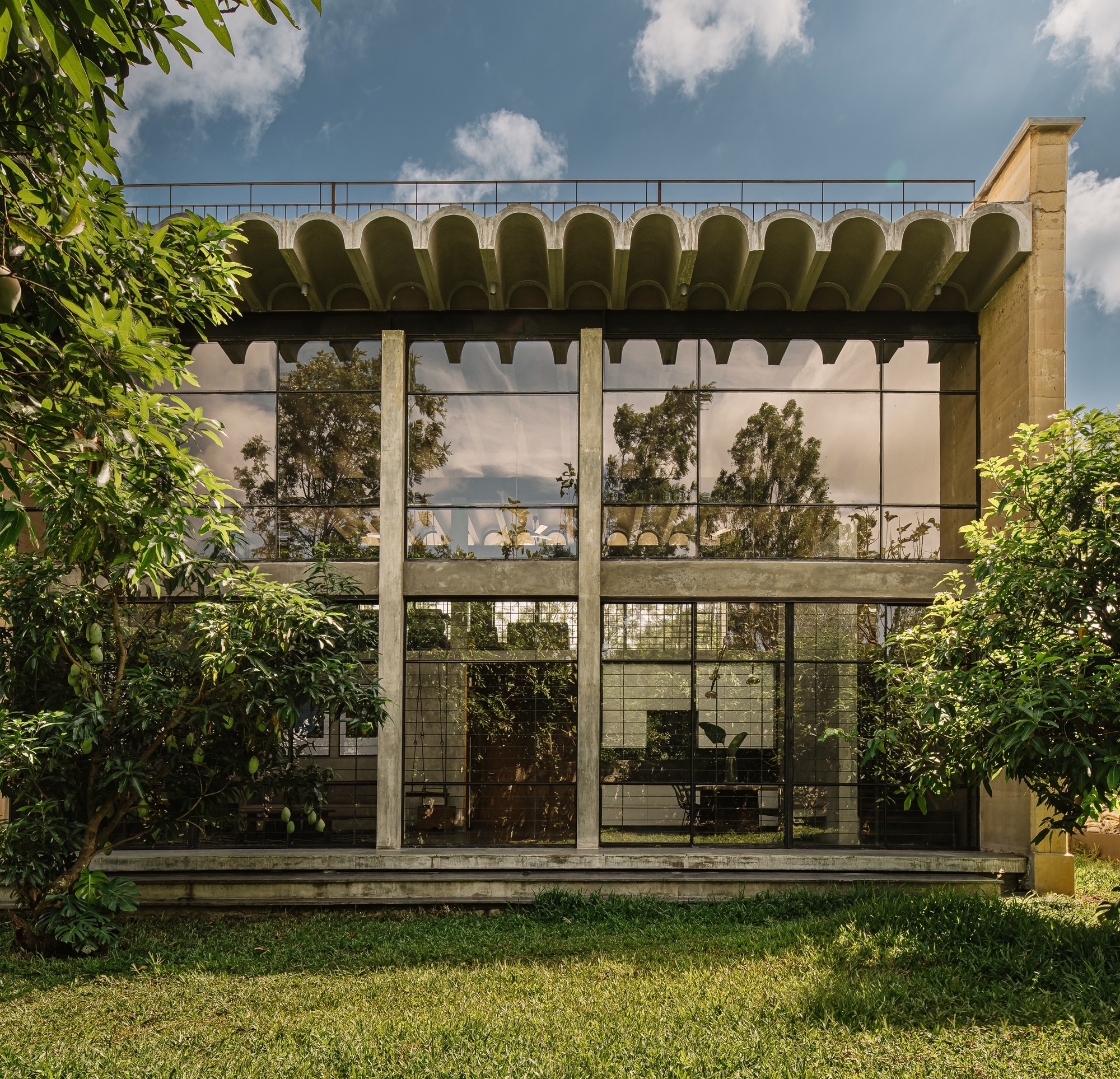 Garden facade with gridded glazing and scalloped concrete canopy framed by lawn and foliage