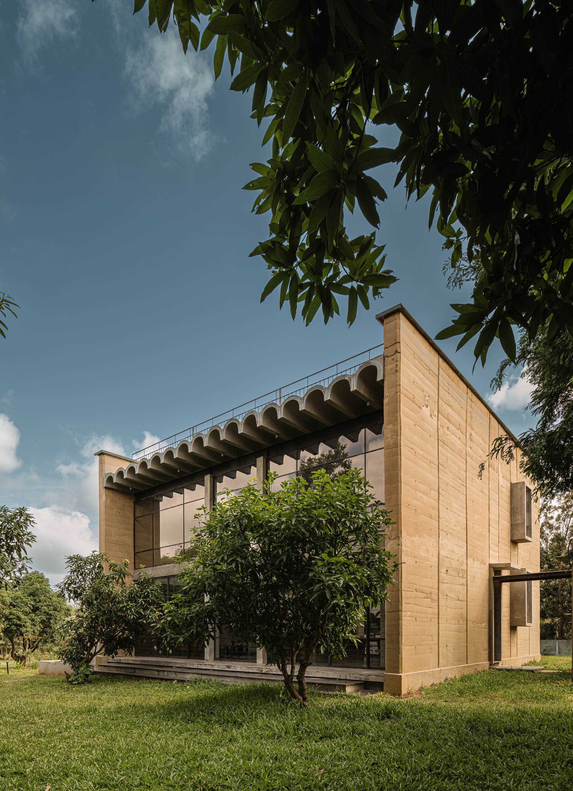 Corner elevation showing rammed earth walls and scalloped canopy amid scattered trees and lawn