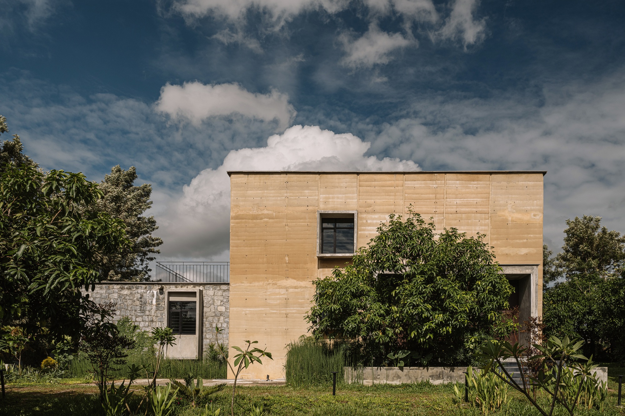 Timber-clad volume with single window rising above a stone base amid tropical vegetation and dramatic clouds