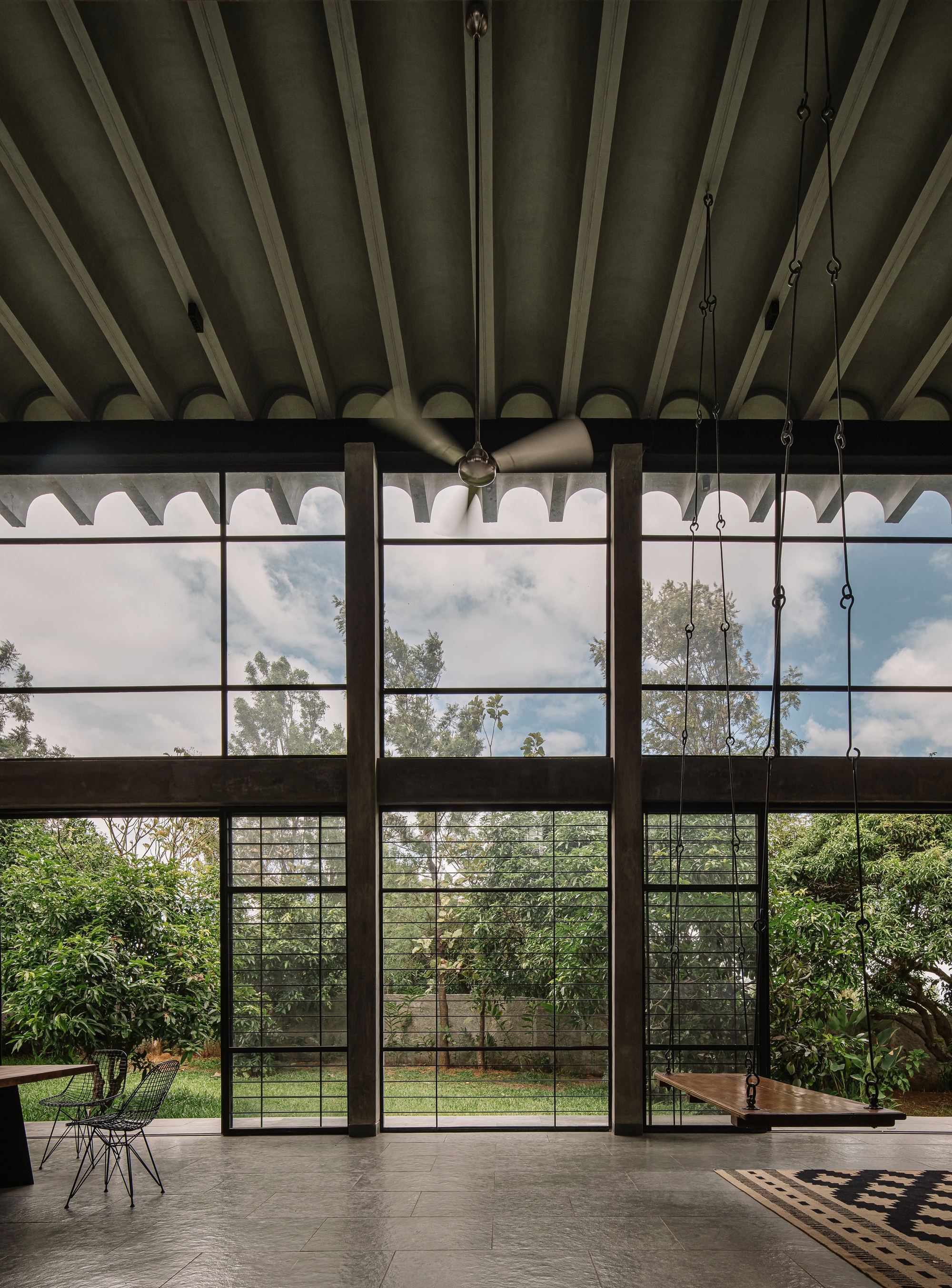 Double-height interior with steel-framed glazing beneath exposed diagonal timber roof trusses and suspended ceiling fan