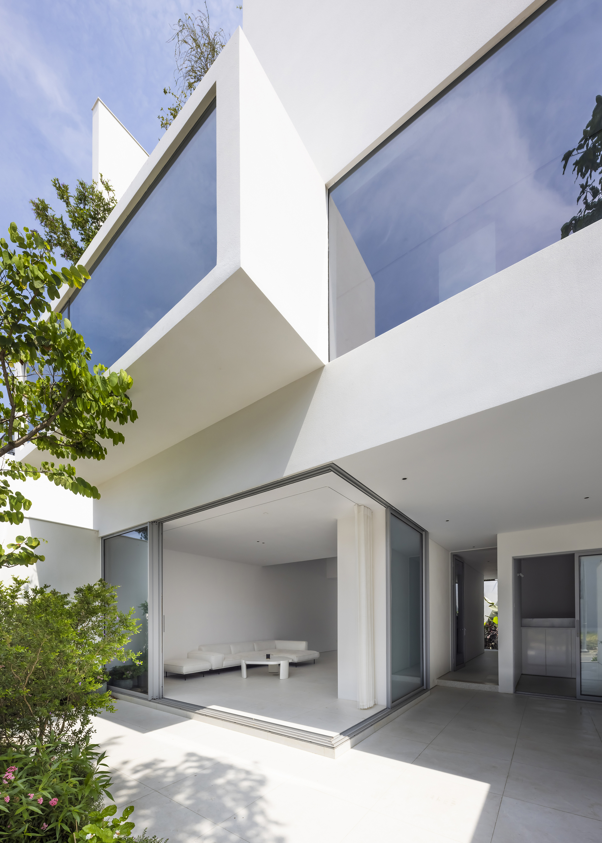 Ground floor terrace with sliding glass doors opening to the interior and trees framing the white volumes above