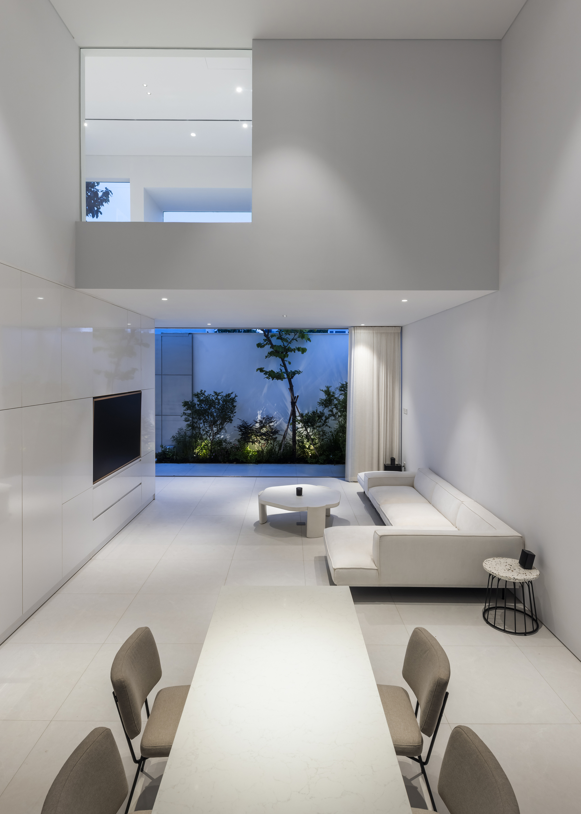 Double-height living room at dusk with suspended mezzanine and garden courtyard view through floor-to-ceiling glazing