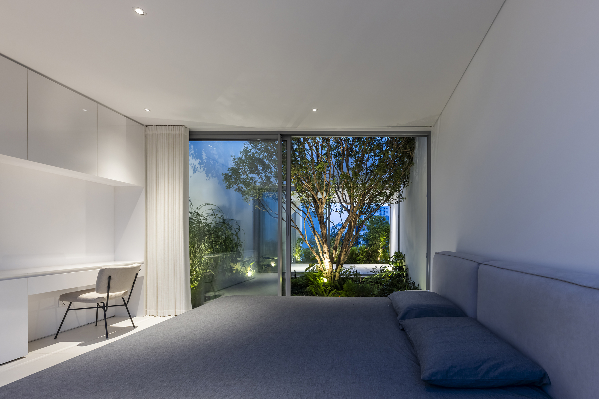 Interior bedroom looking through glass doors to a courtyard with an uplighted tree at dusk