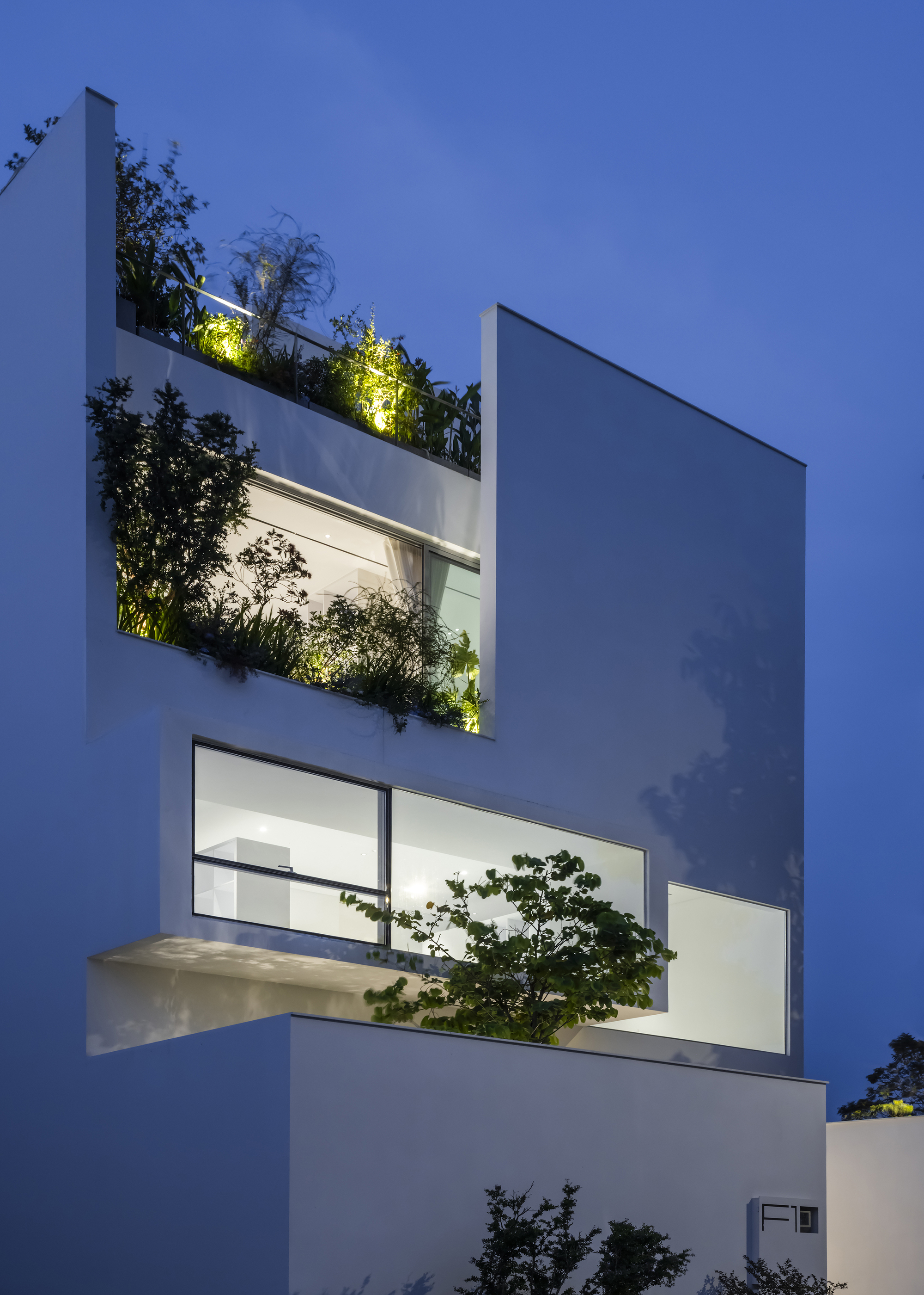 White facade with recessed planted terraces and illuminated greenery against a darkening evening sky