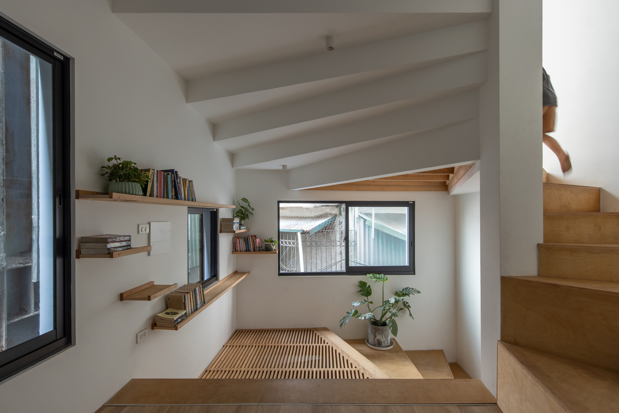 Interior reading nook with timber platform, slatted floor, floating shelves, and person ascending the staircase