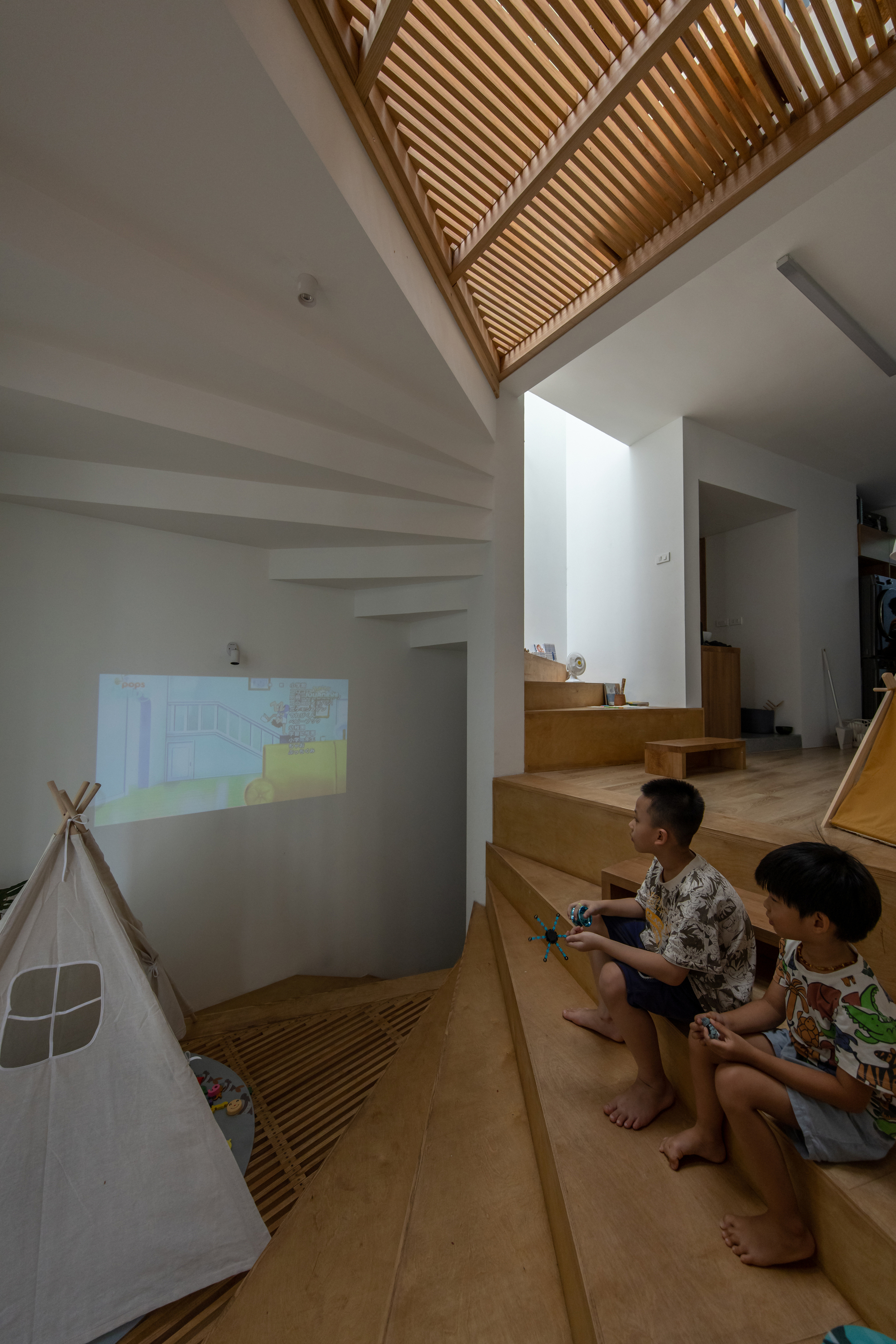 Double-height space with slatted timber ceiling and two children seated on the stepped platform